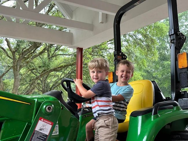 Two children sit on a green tractor under a porch, smiling at the camera.