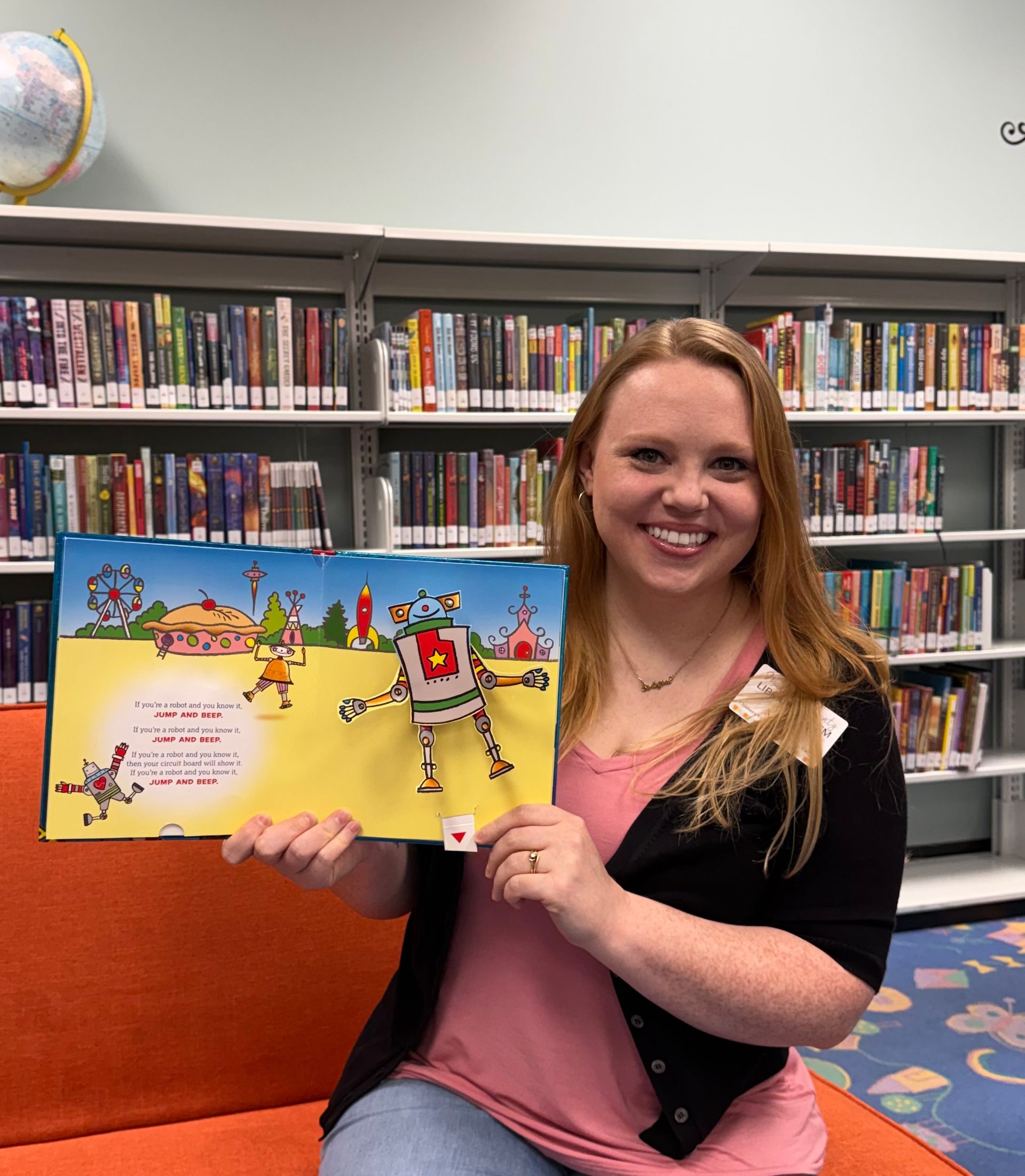 Woman smiling in a library, holding up an open children’s book with bright illustrations.