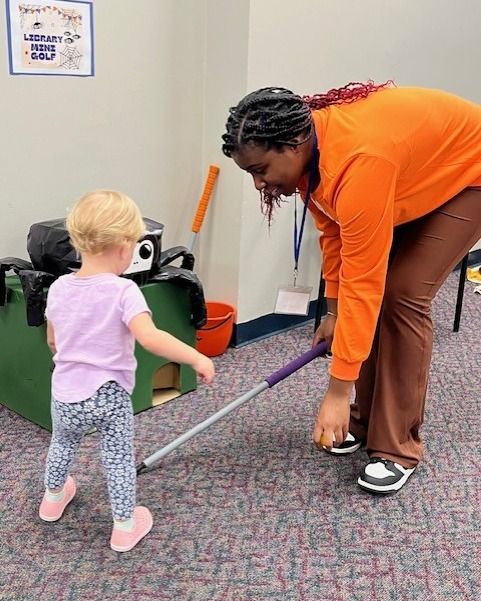 Adult and toddler touching a long measuring stick on a carpeted floor in a classroom.