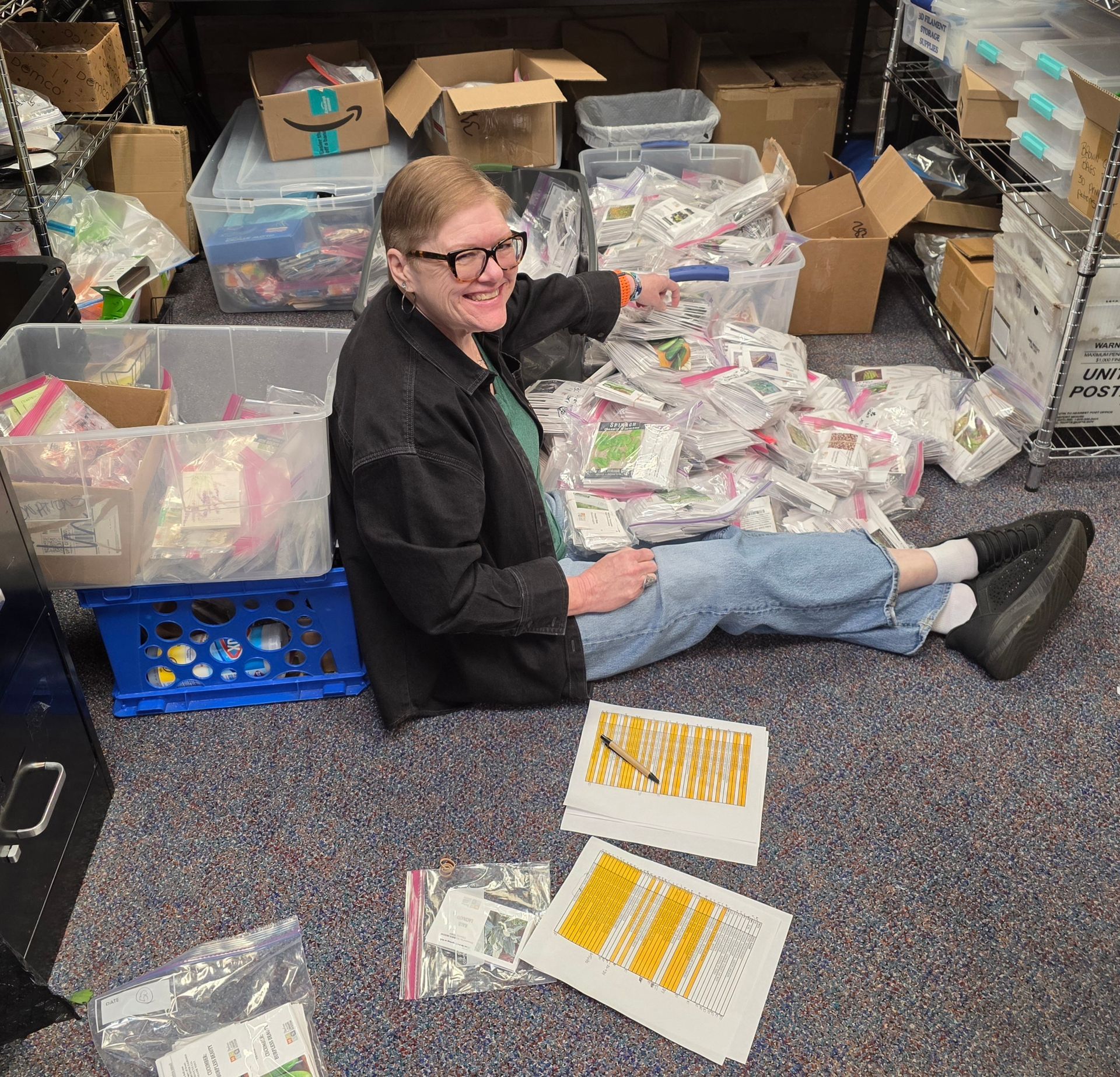 Person sitting on the floor among boxes and supplies, smiling in a cluttered storage room.