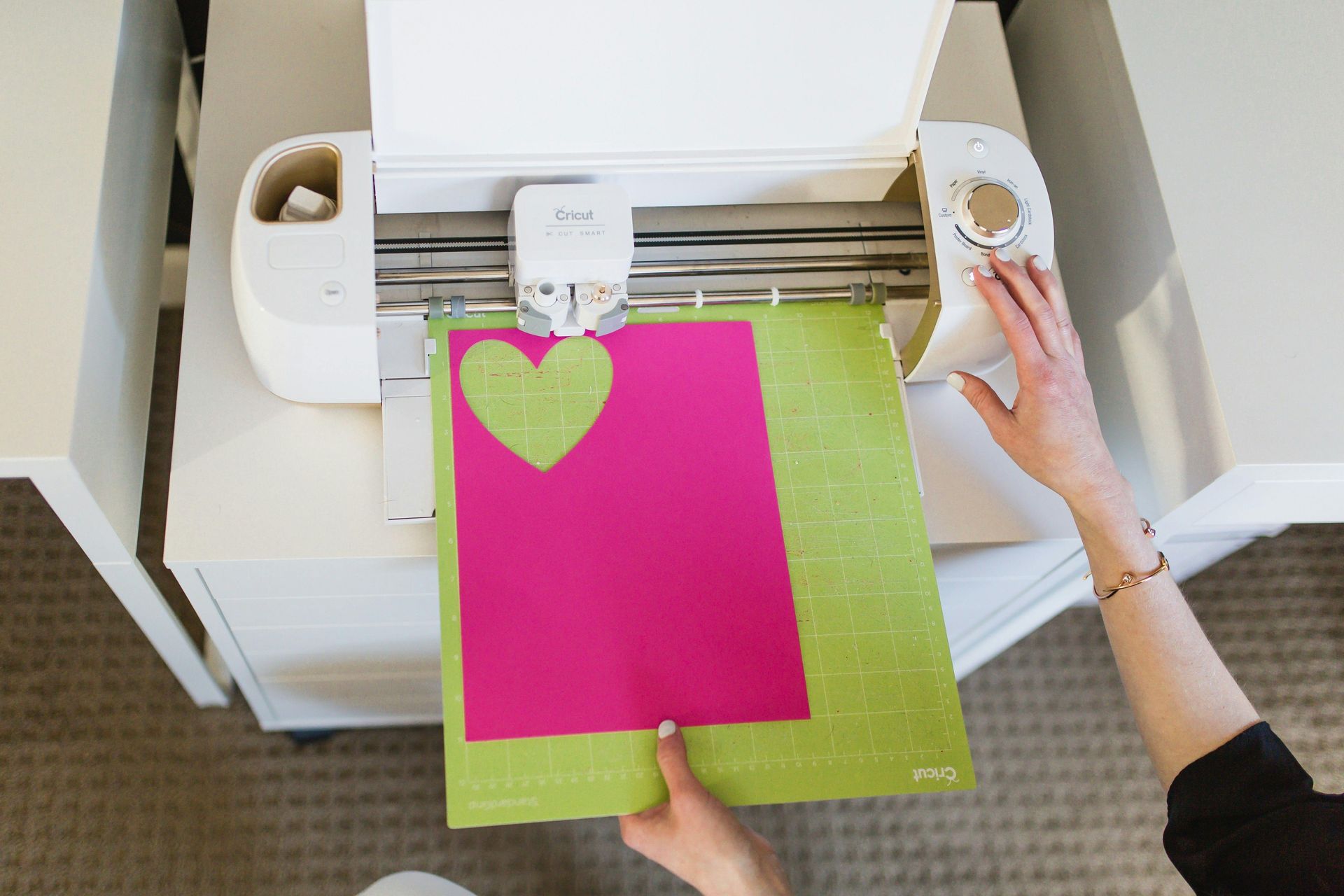 Hands feed pink-and-lime paper into a cutting machine in a craft workspace.