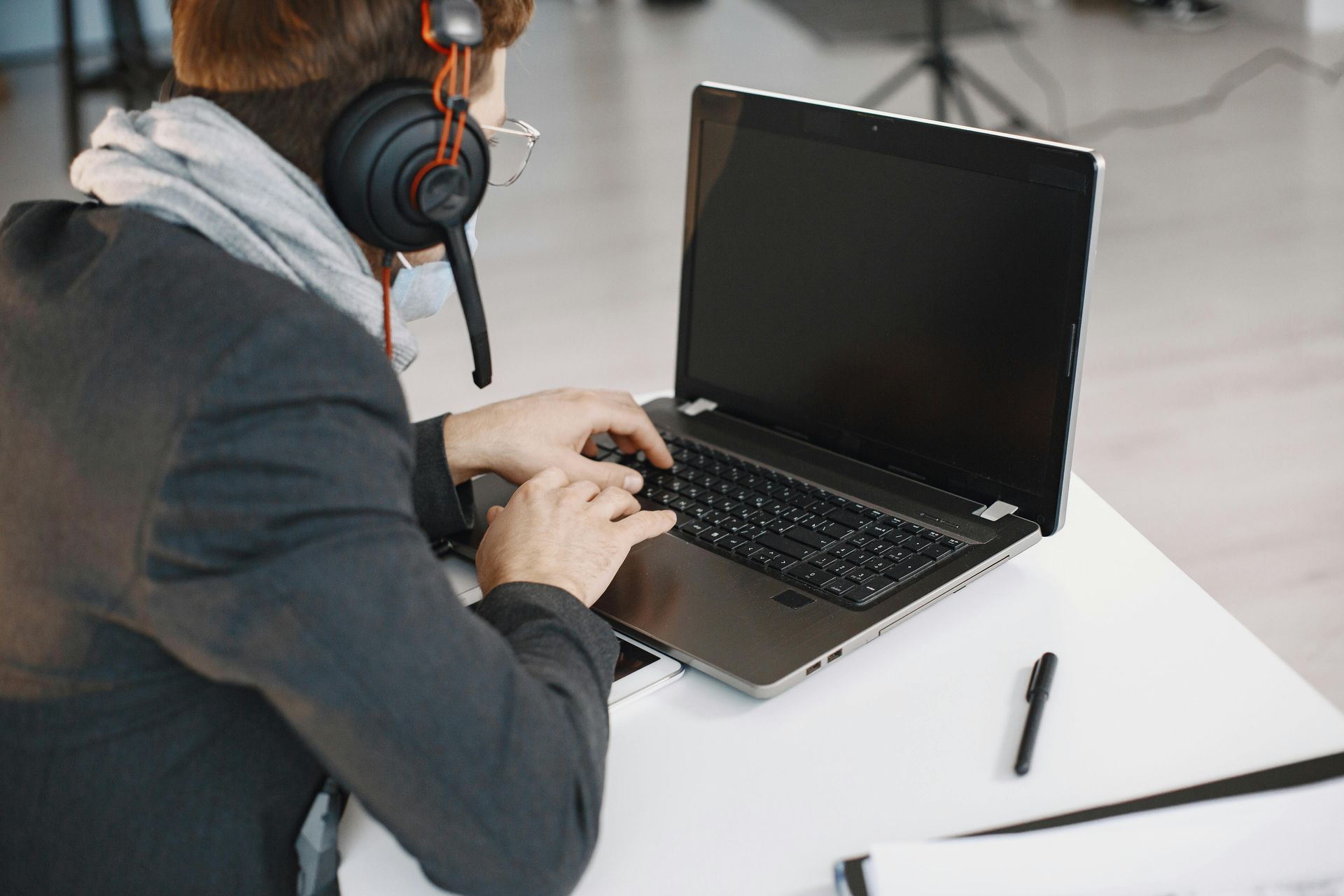 Person wearing headphones typing on a laptop at a white desk with a pen nearby