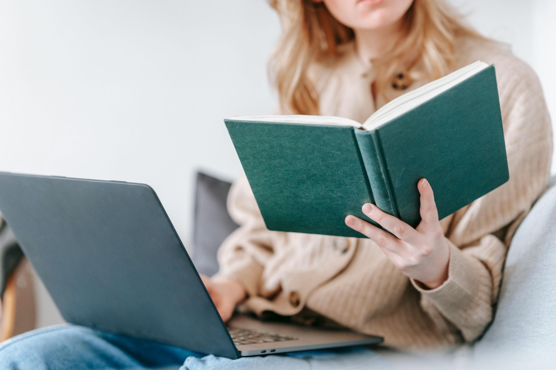 Person reading a green book beside a laptop on a couch