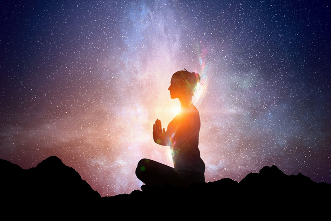 Silhouette of a person meditating against a glowing starry sky and rocky foreground