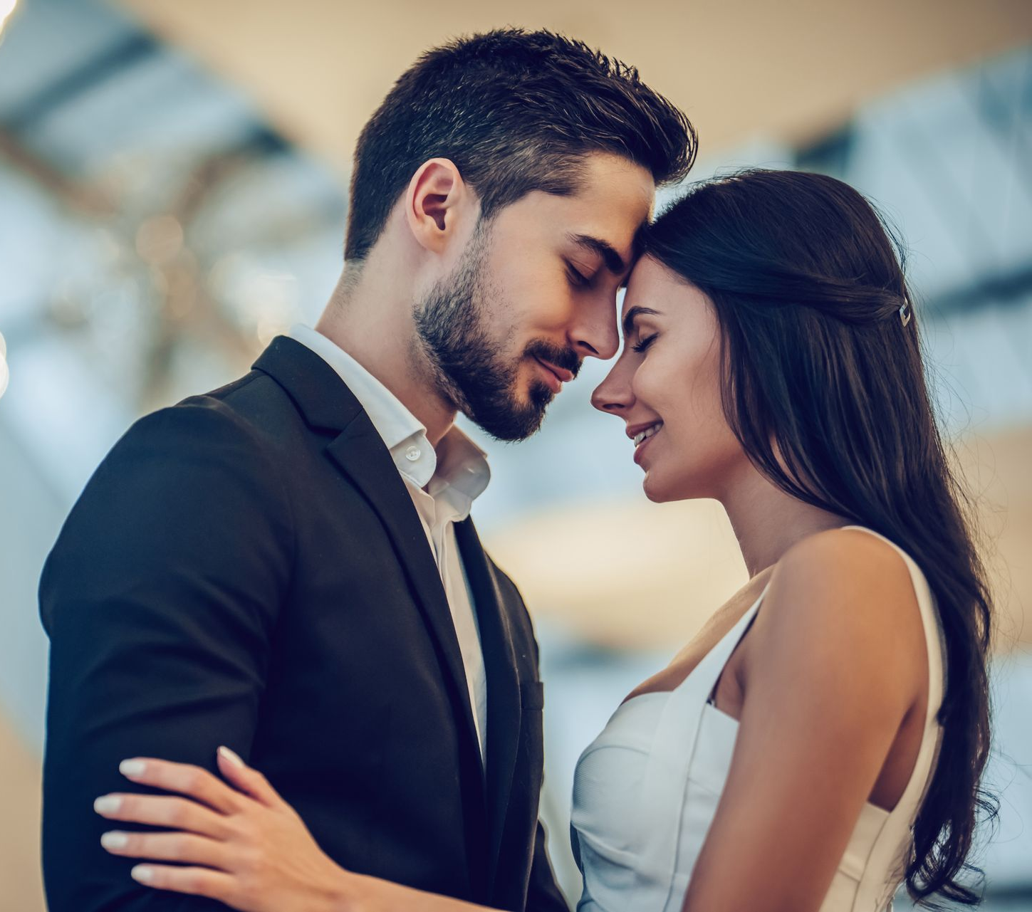 Couple embracing and smiling closely, dressed formally in a softly blurred indoor setting