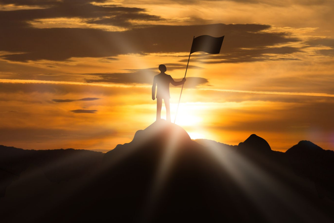Silhouetted figure on a peak holding a flag at sunset, with glowing light and clouds behind.