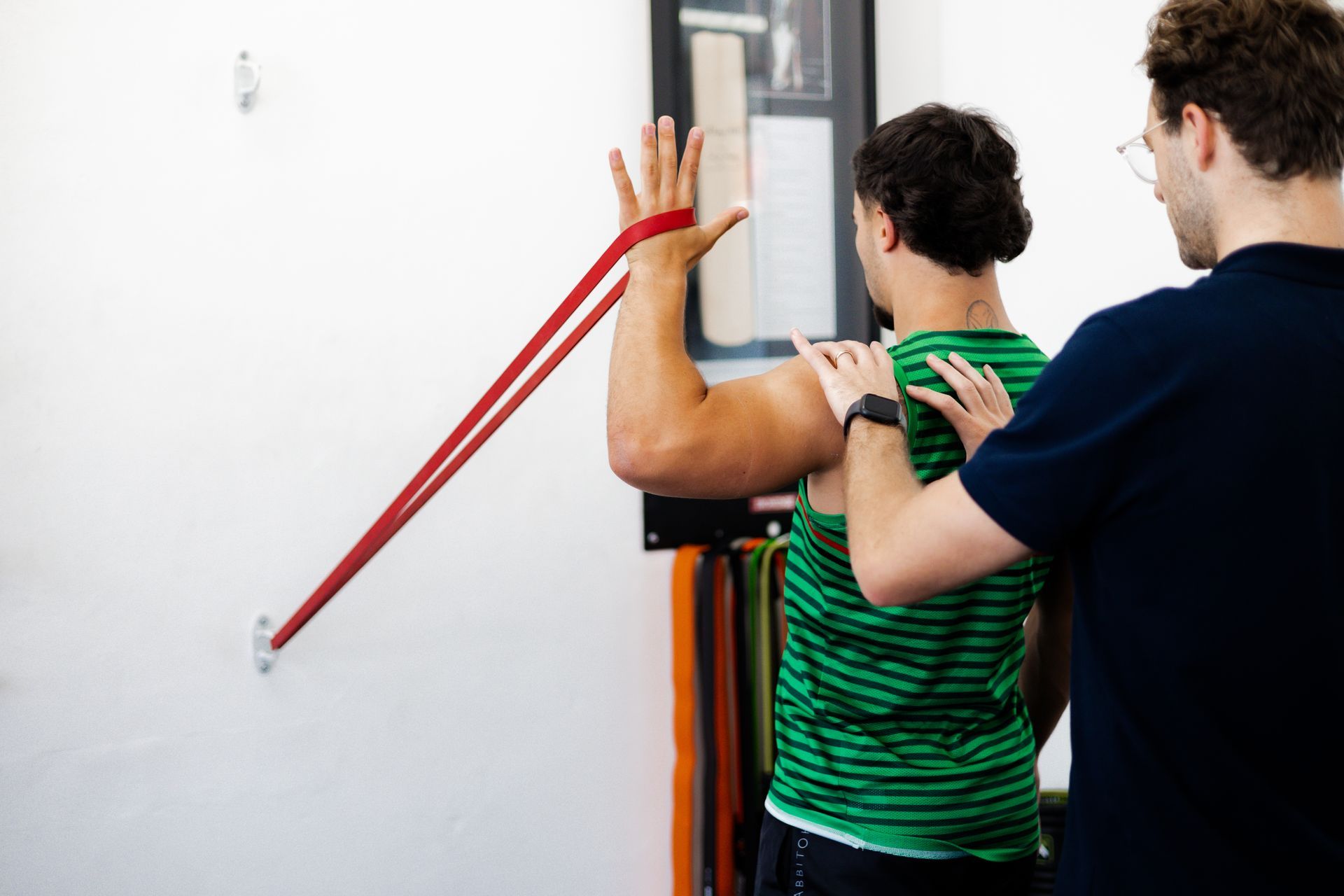 Man doing shoulder exercise with red resistance band, aided by an instructor.