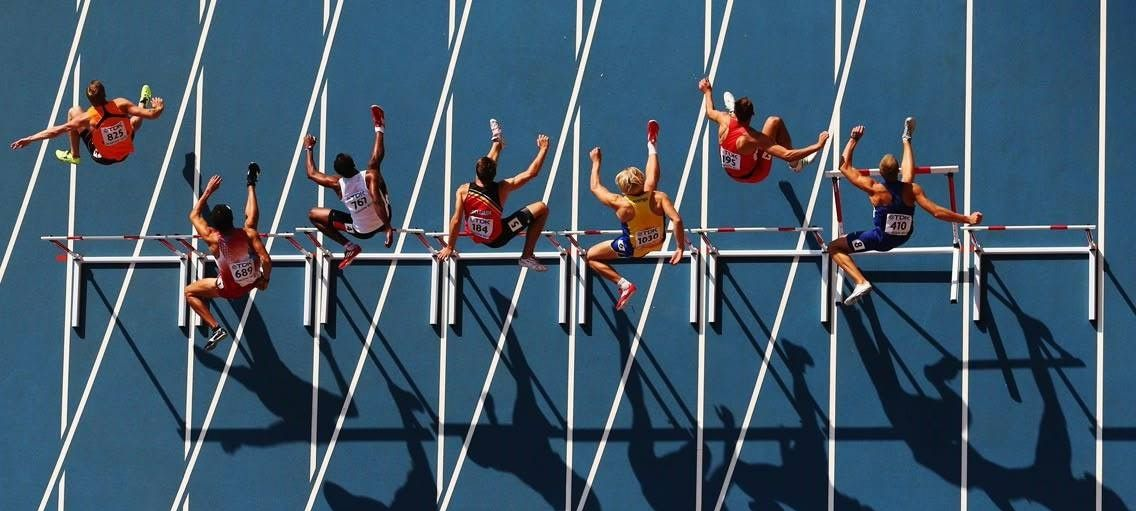 Athletes leap over hurdles on a blue track. Shadows stretch, sunlight strong.