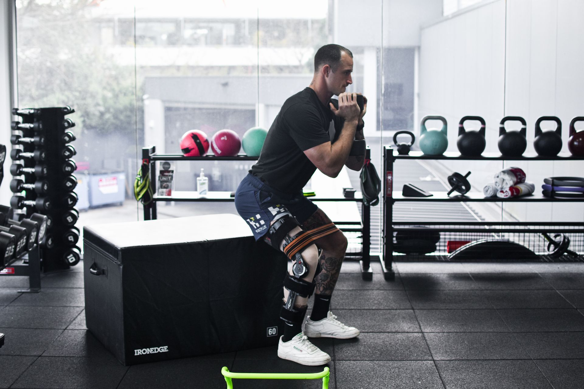 Man doing a squat with a dumbbell, using a box and resistance band in a gym.
