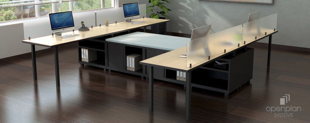 Office desks with plexiglass dividers. Monitors sit atop light-colored wood tables with black legs.