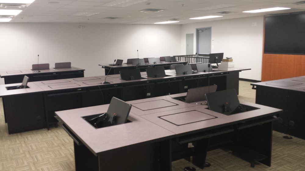 A classroom with several dark brown desks, each with a computer. White walls and beige carpet.