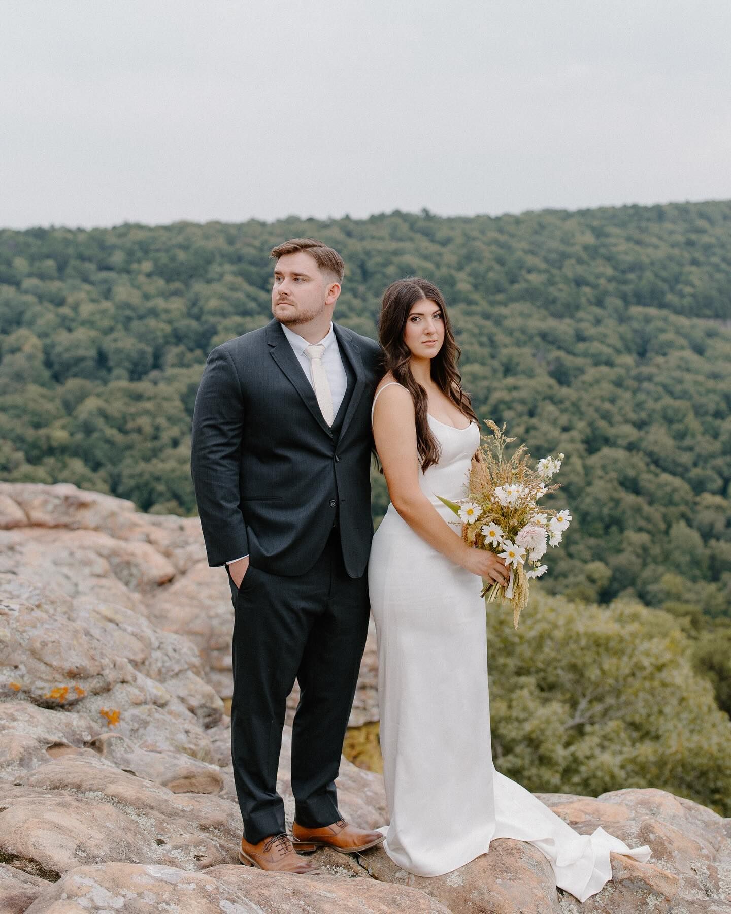 A bride and groom are standing on top of a rocky hill.