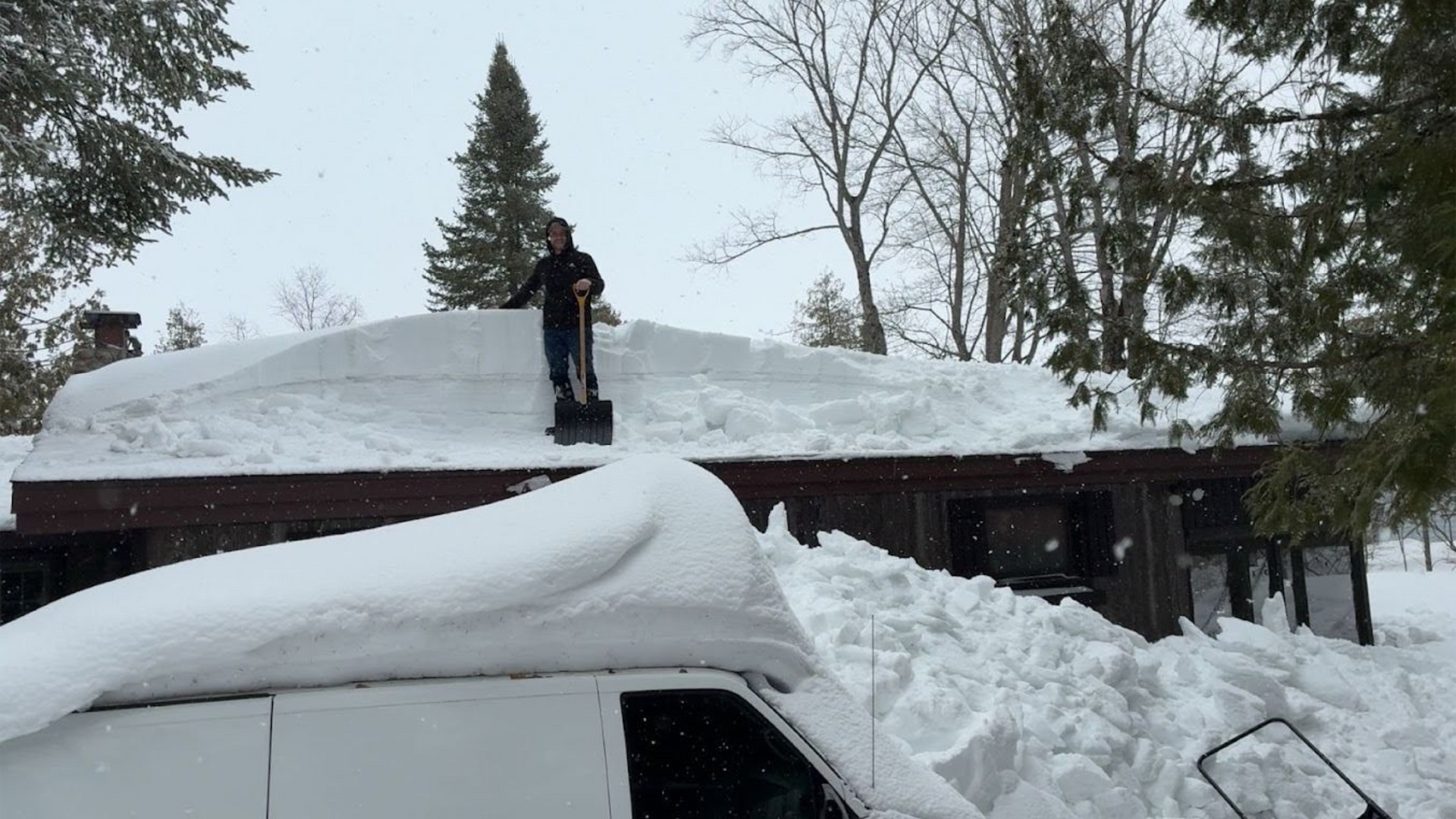 Person shoveling heavy snow off a roof above a snow-covered vehicle and driveway