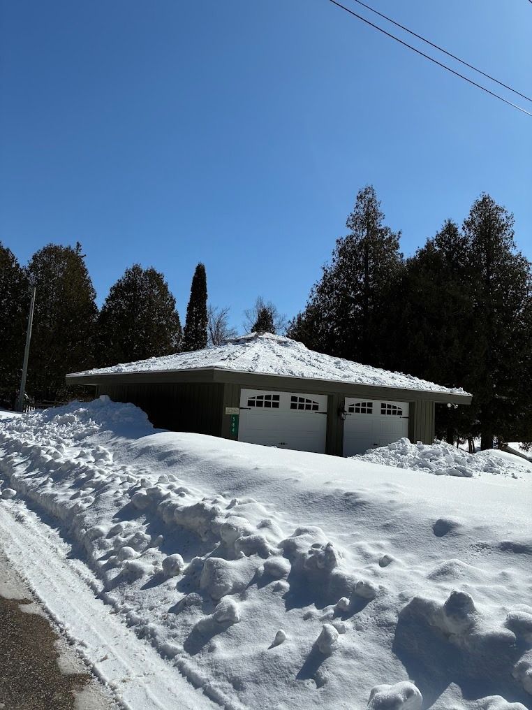 Snow-covered house with double garage under a clear blue sky, surrounded by trees.