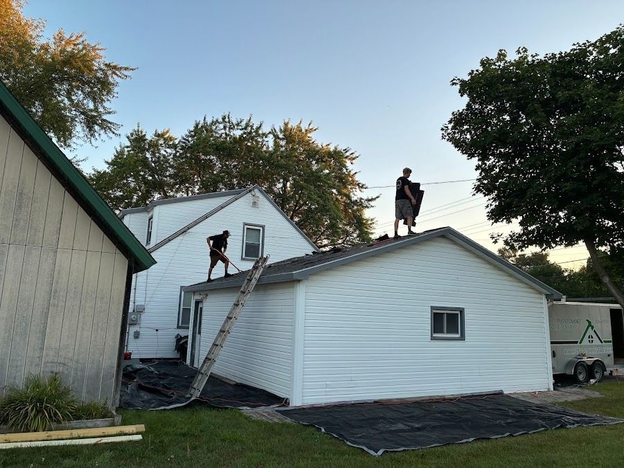 Two workers on a ladder repairing a white house roof beside a green-sided building.
