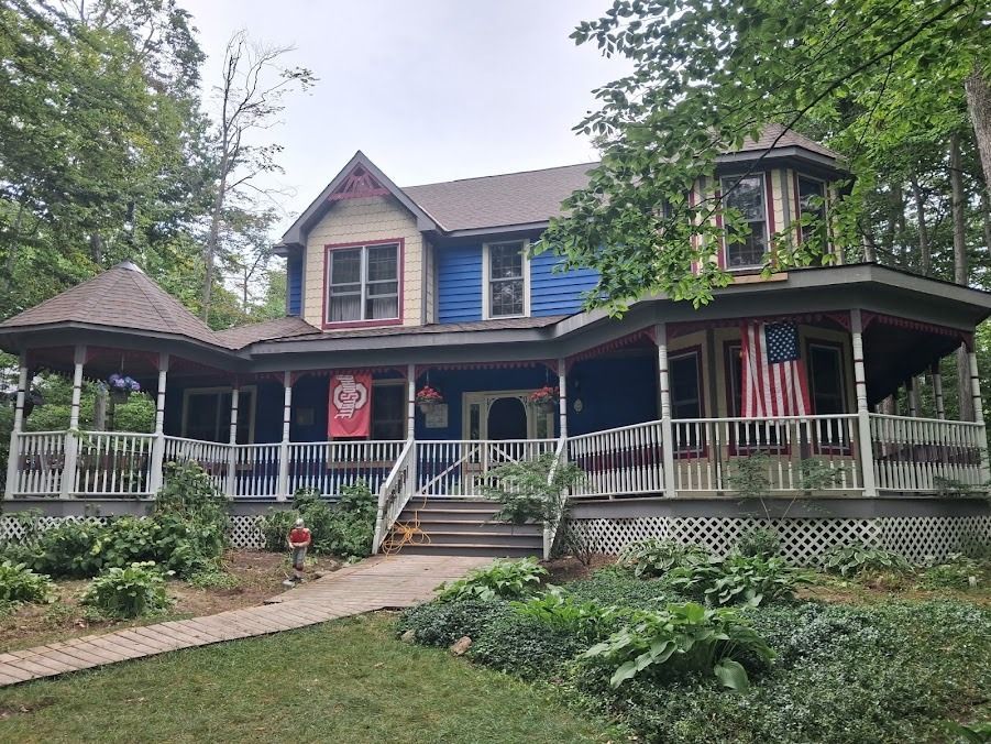 Victorian house with wraparound porch, white railings, blue shutters, and American flags in a leafy yard