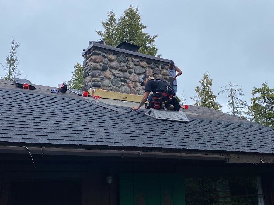Roofers working on a shingled roof beside a stone chimney on a cabin