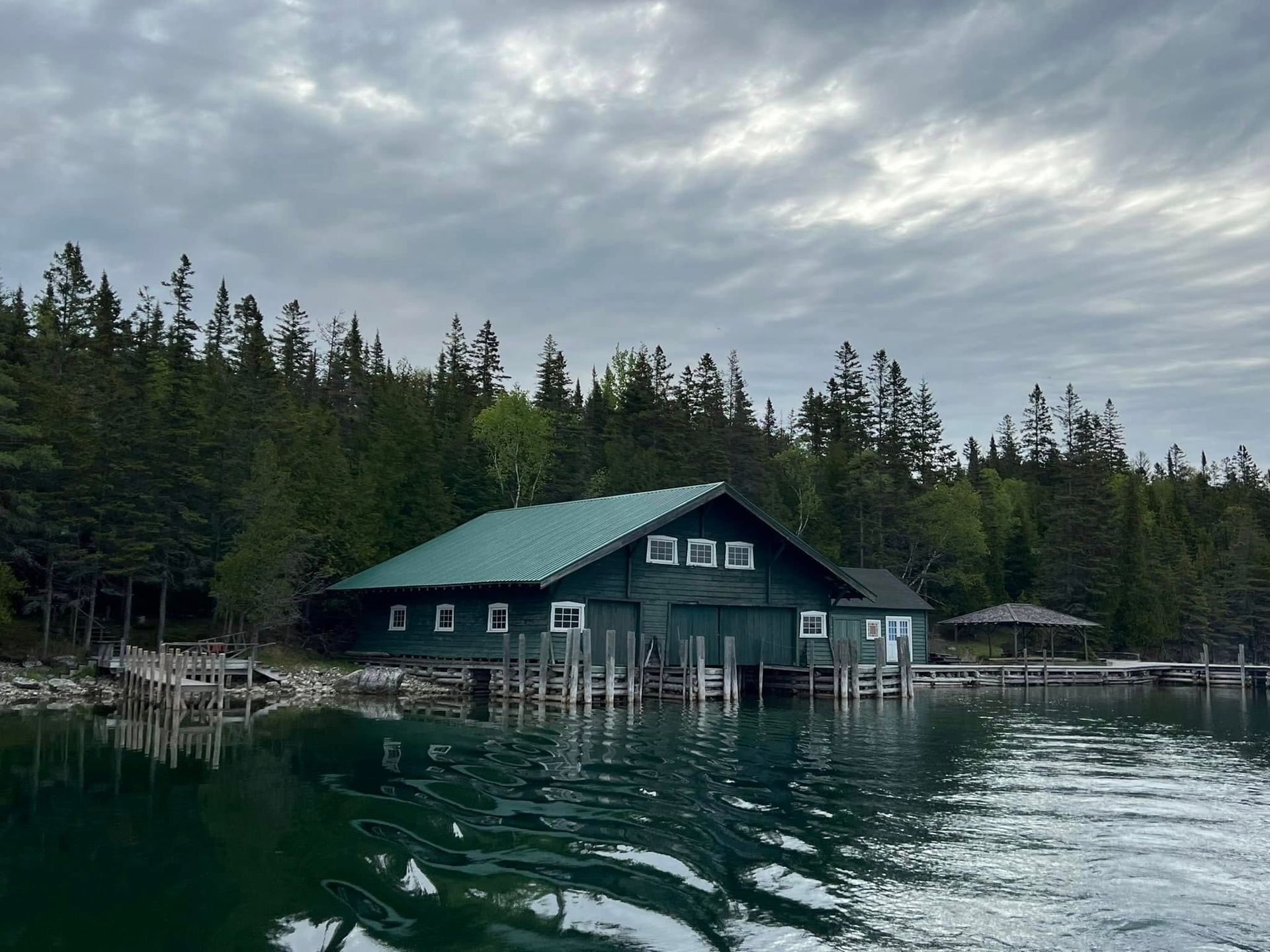 Dark green lakeside boathouse with dock, surrounded by pine trees under a cloudy sky