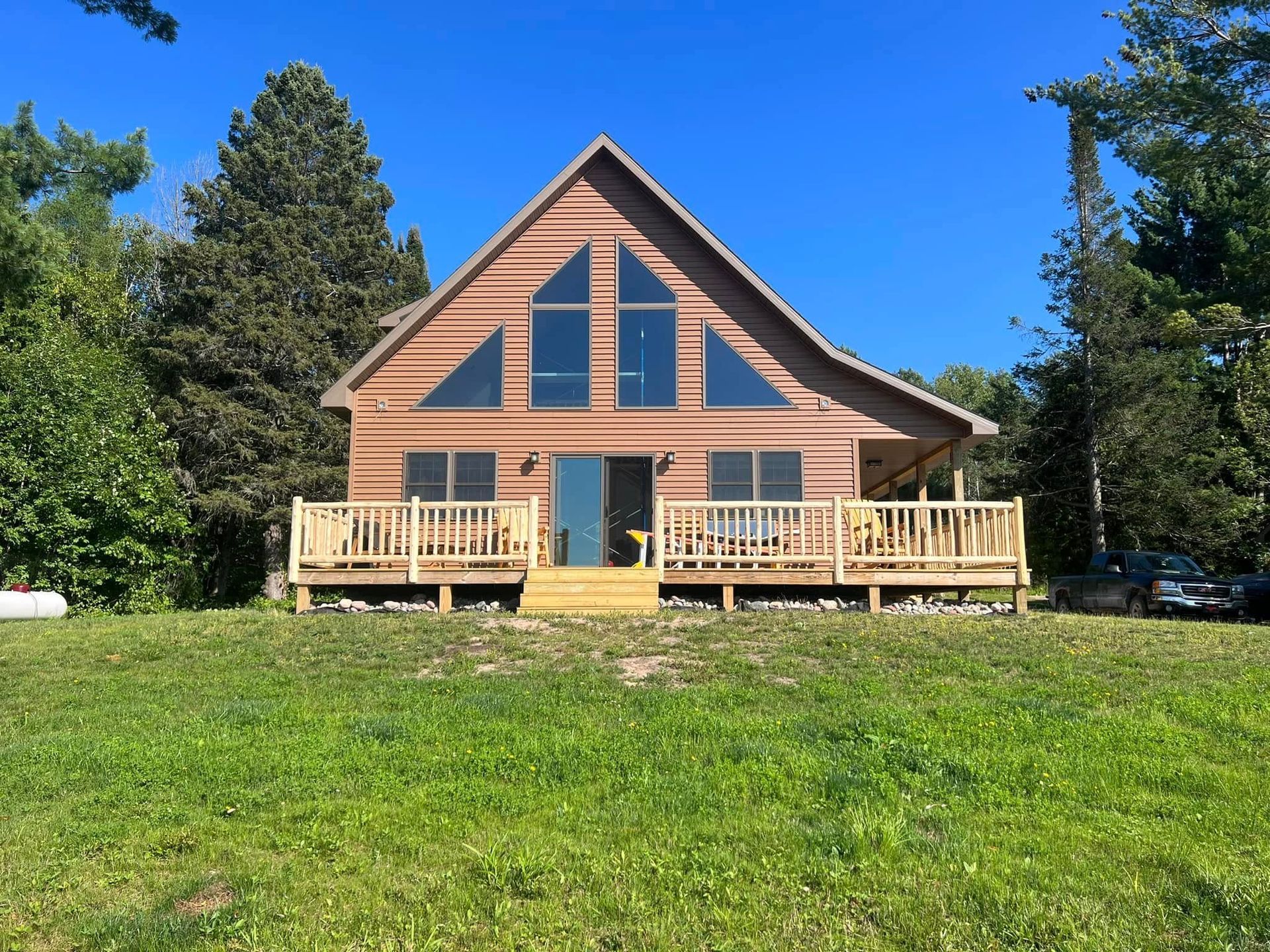 Wooden cabin with large windows and front deck on a sunny green lawn, surrounded by trees.
