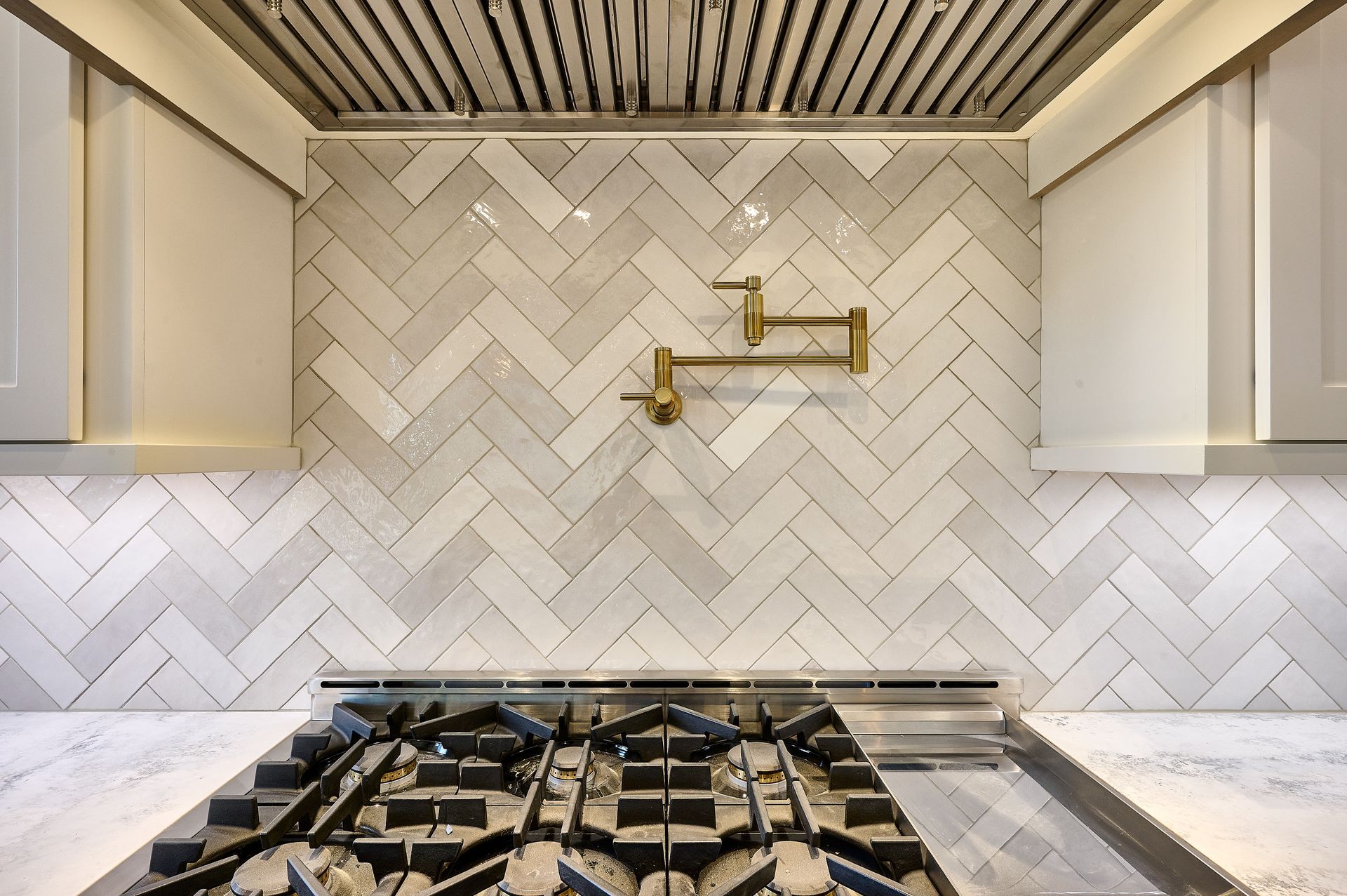 White herringbone tile backsplash with gold faucet above gas stovetop in a kitchen.