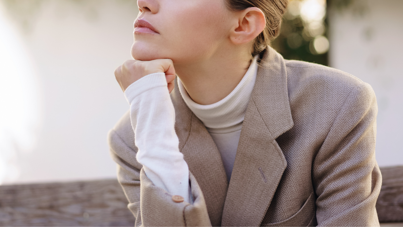 Woman in tan blazer and white turtleneck, resting chin on hand, looking upward.