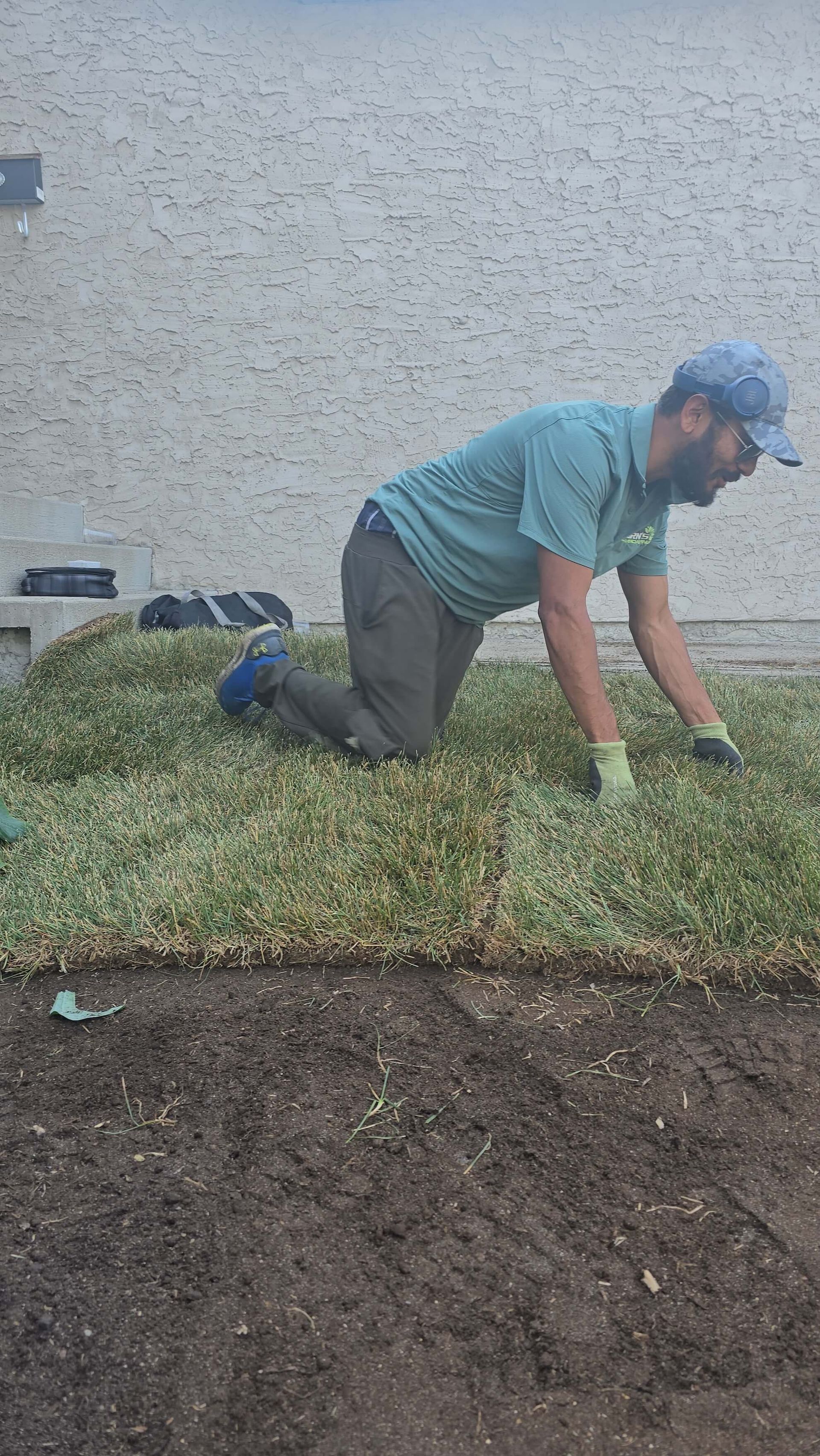 Man laying sod on a patch of soil, kneeling with a cap, gloves, and a green shirt near a wall.