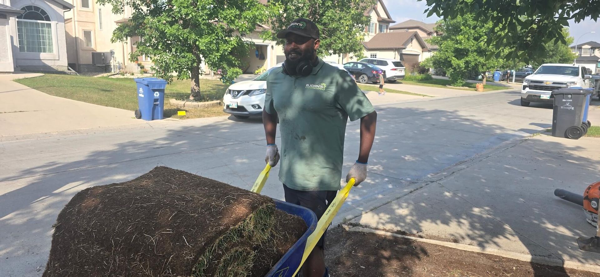 A person pushing a wheelbarrow filled with dirt, on a residential street.