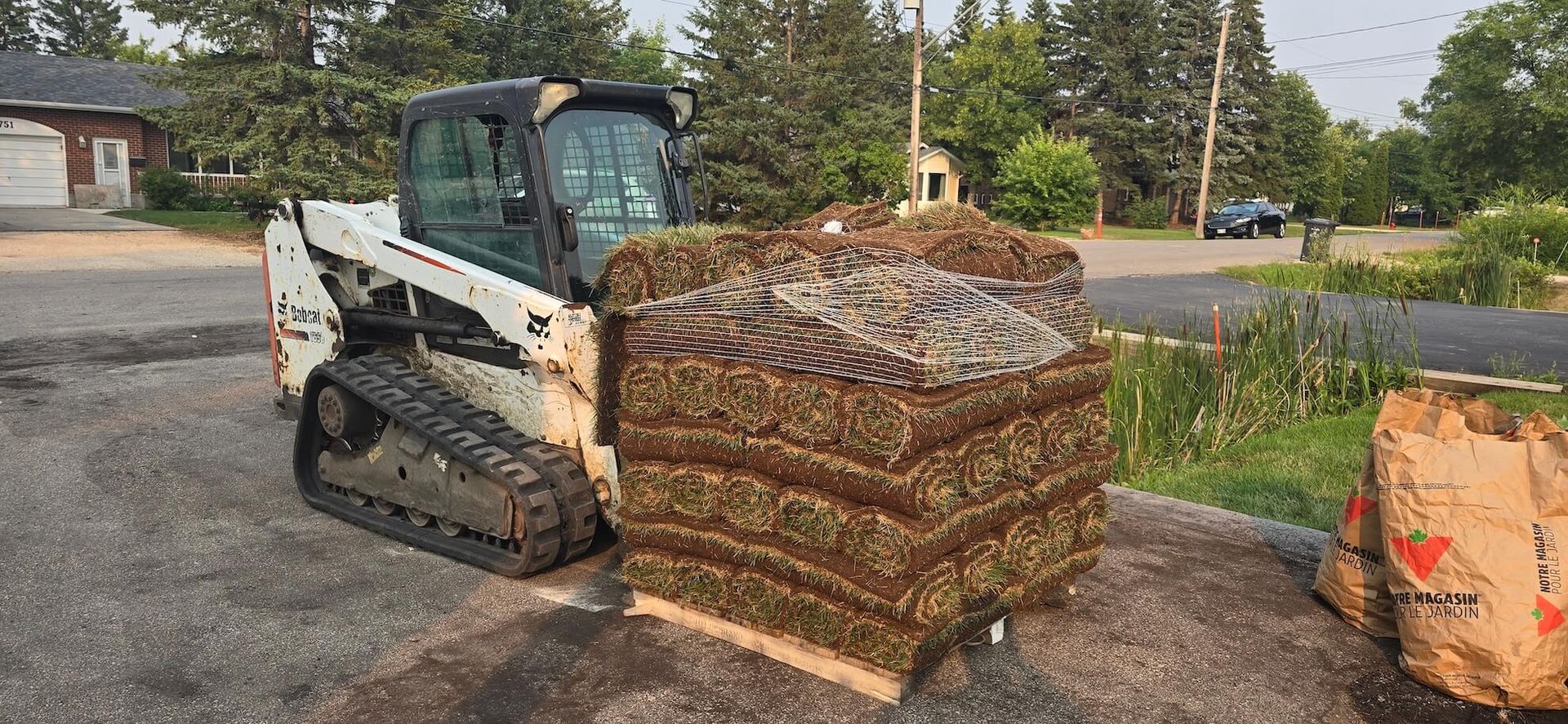 A Bobcat skid steer next to stacked sod on a paved surface.