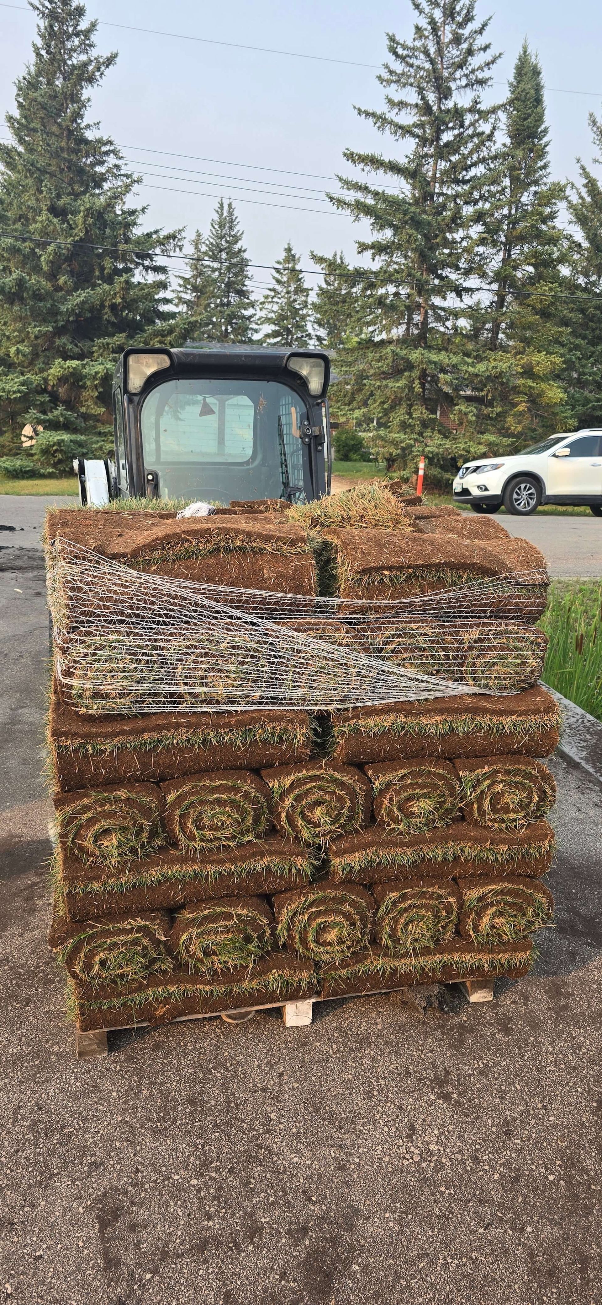 A stack of sod squares on a pallet, with a skid steer and trees in the background.