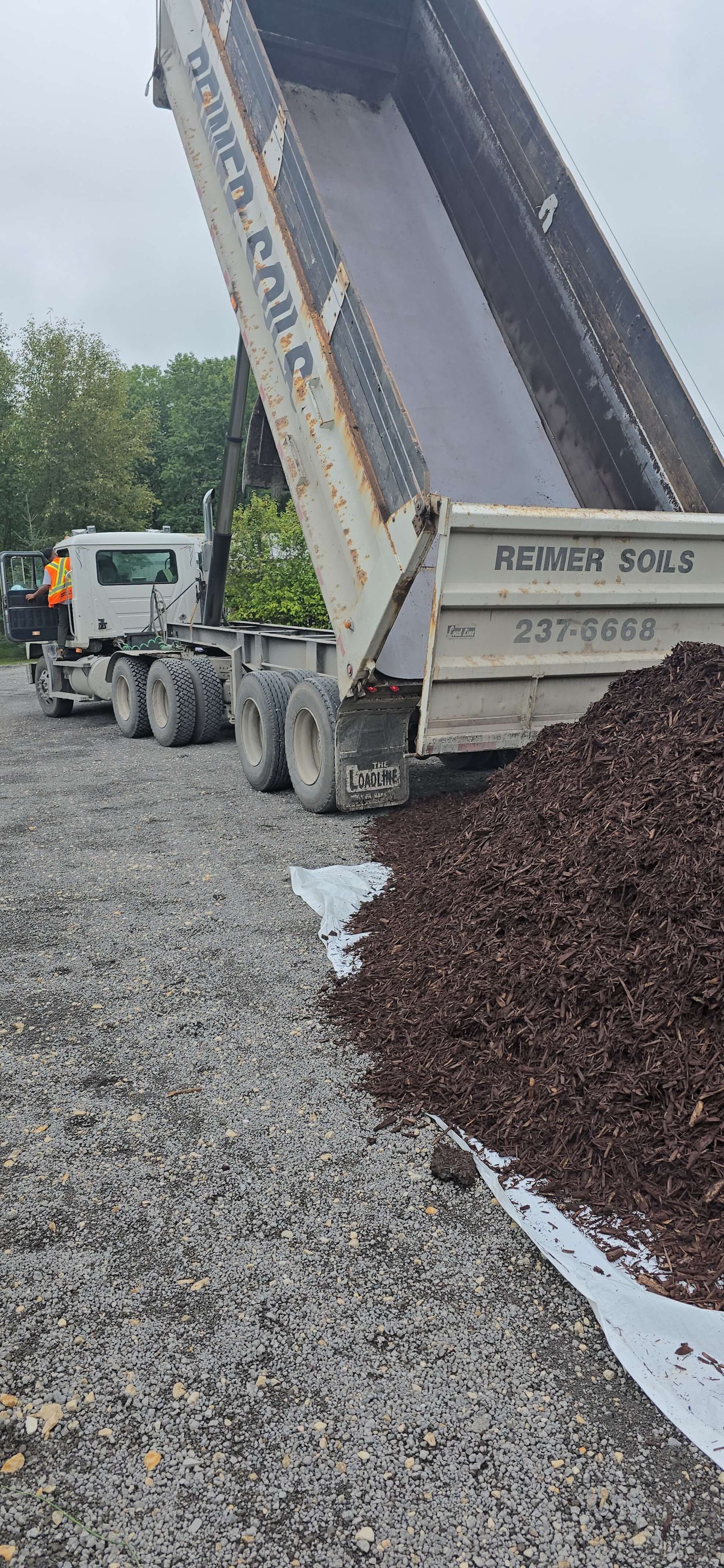 A dump truck pouring dark gravel onto a bed of lighter gravel on a cloudy day.