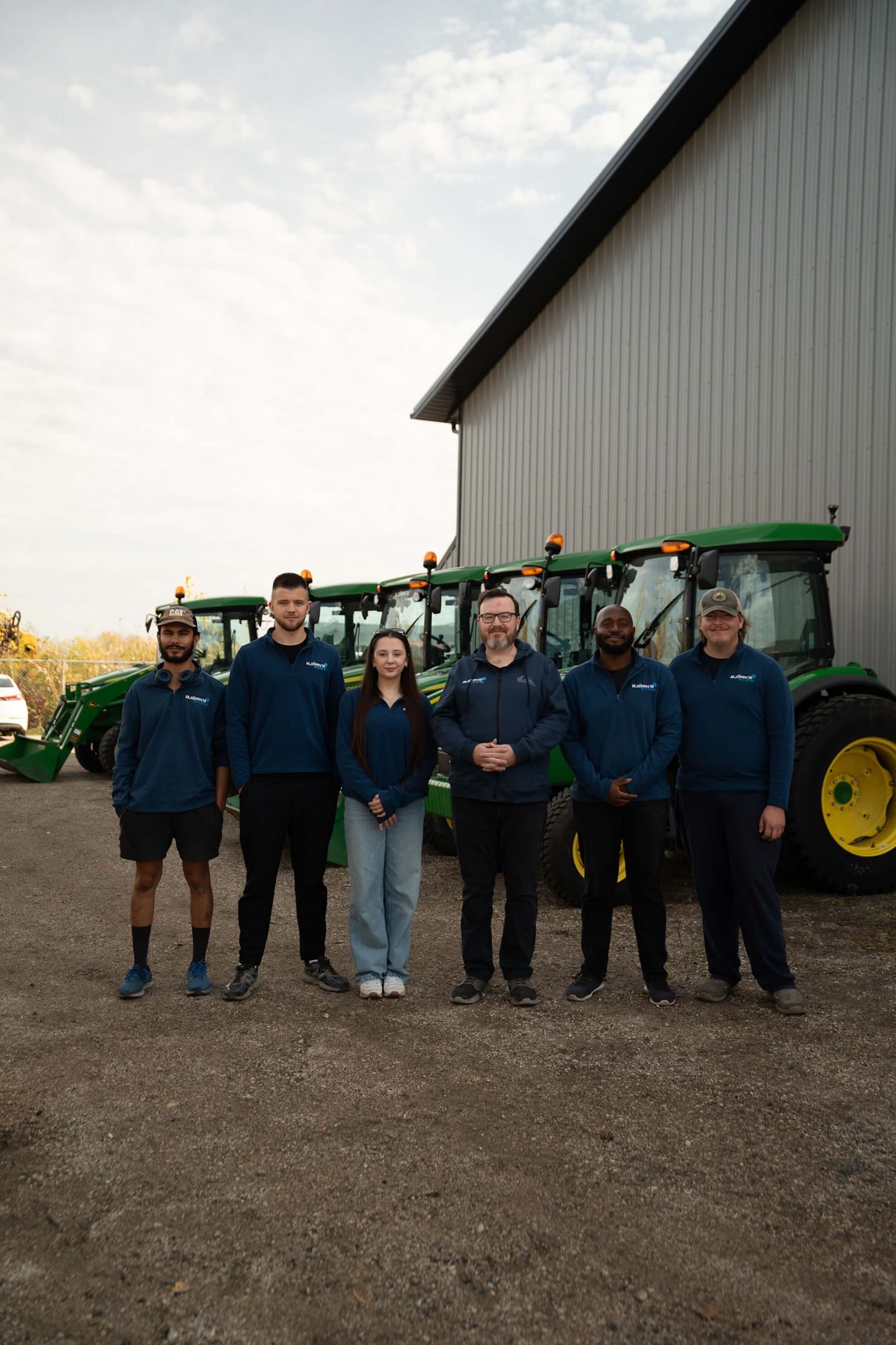 Group of six people in blue jackets stand in front of green tractors and a gray building.