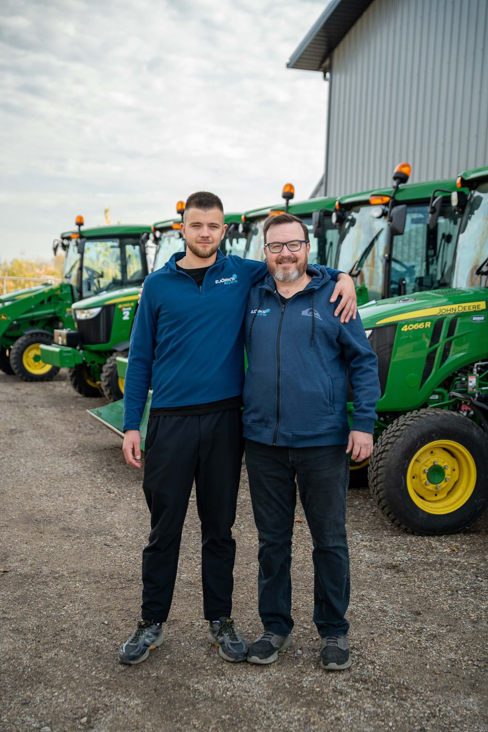 Two men stand by green tractors; one has arm around the other. Exterior; cloudy day.