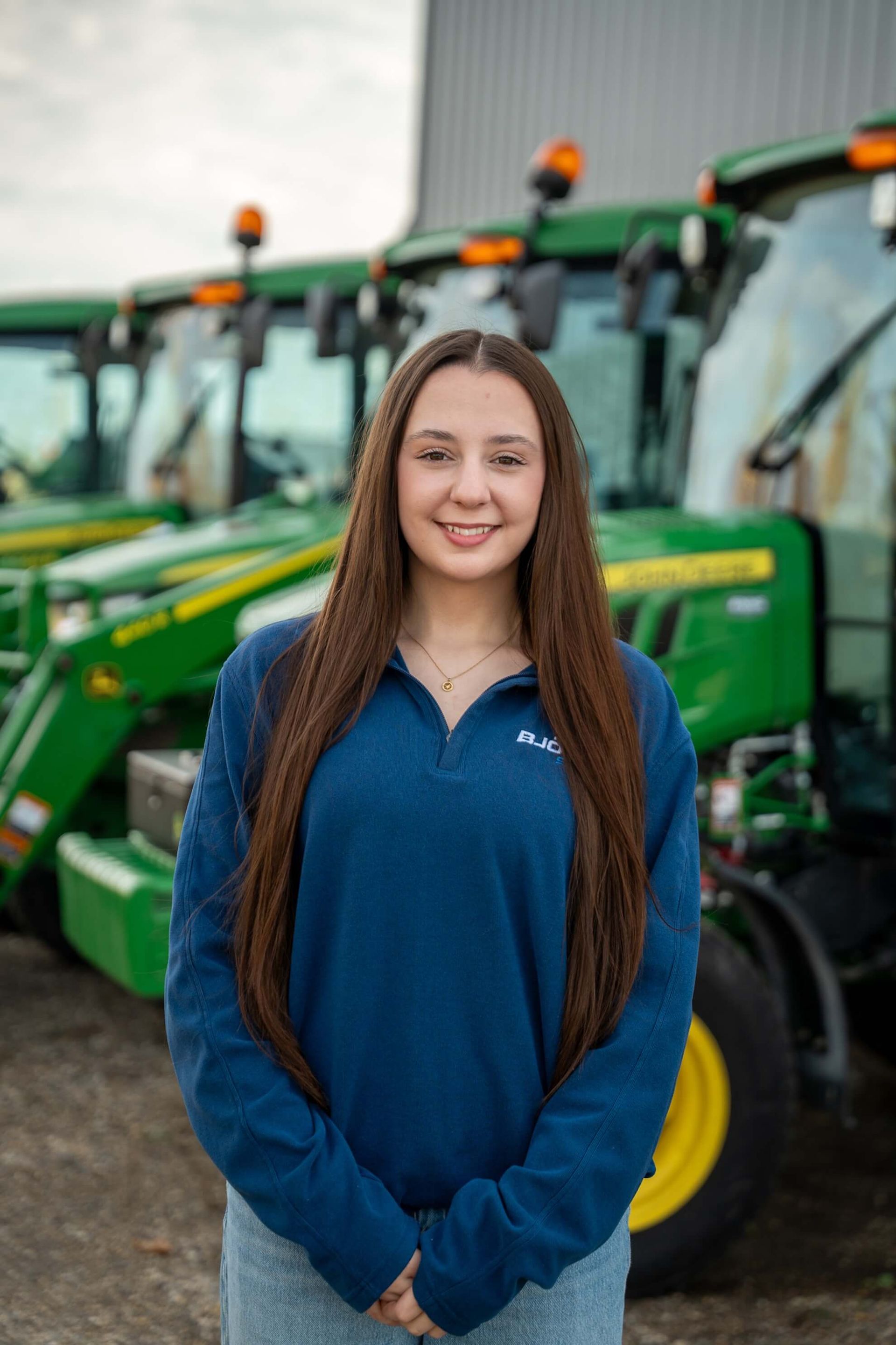 Woman in blue shirt smiles in front of green tractors.
