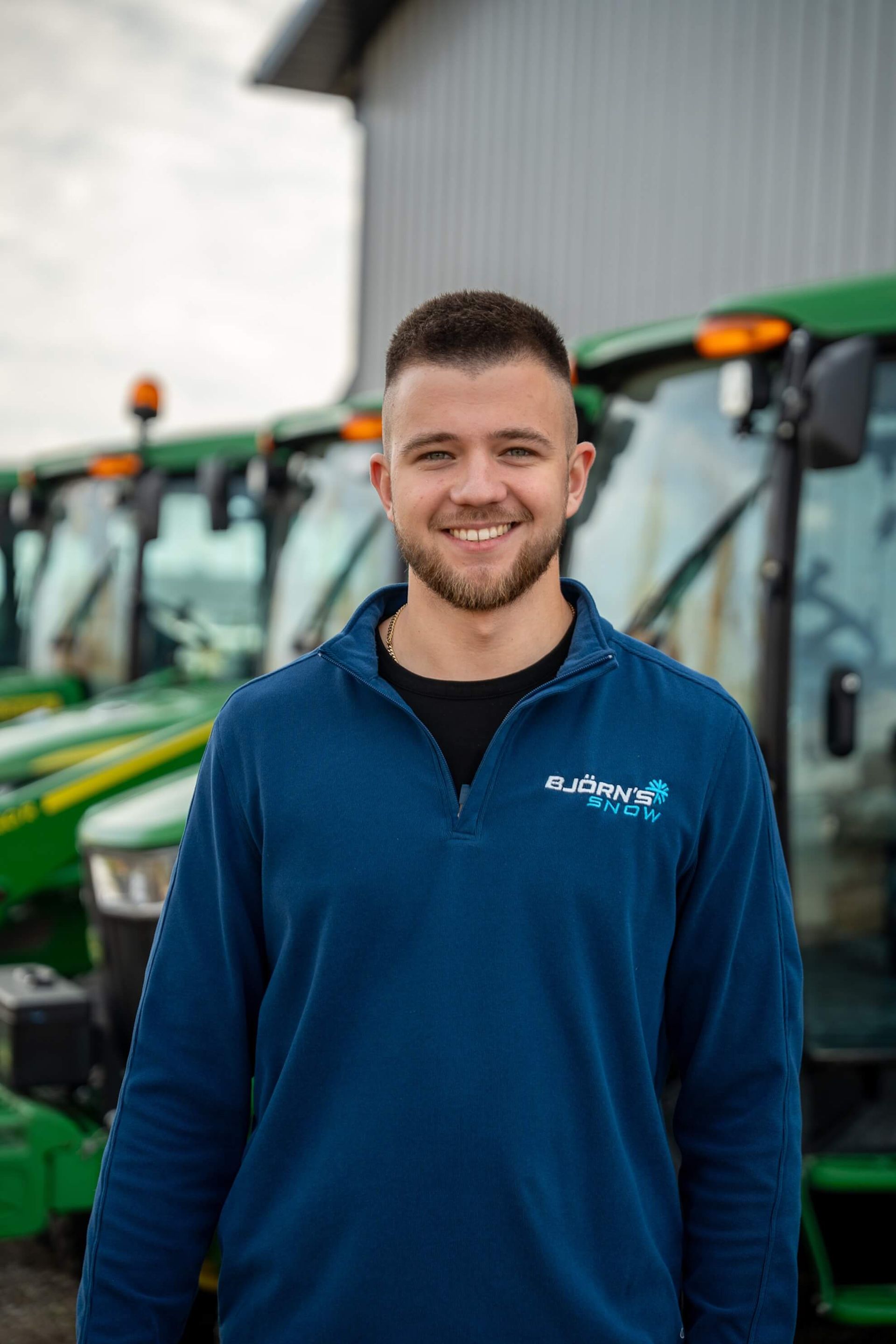 Man smiling, wearing a blue pullover, standing in front of green tractors.