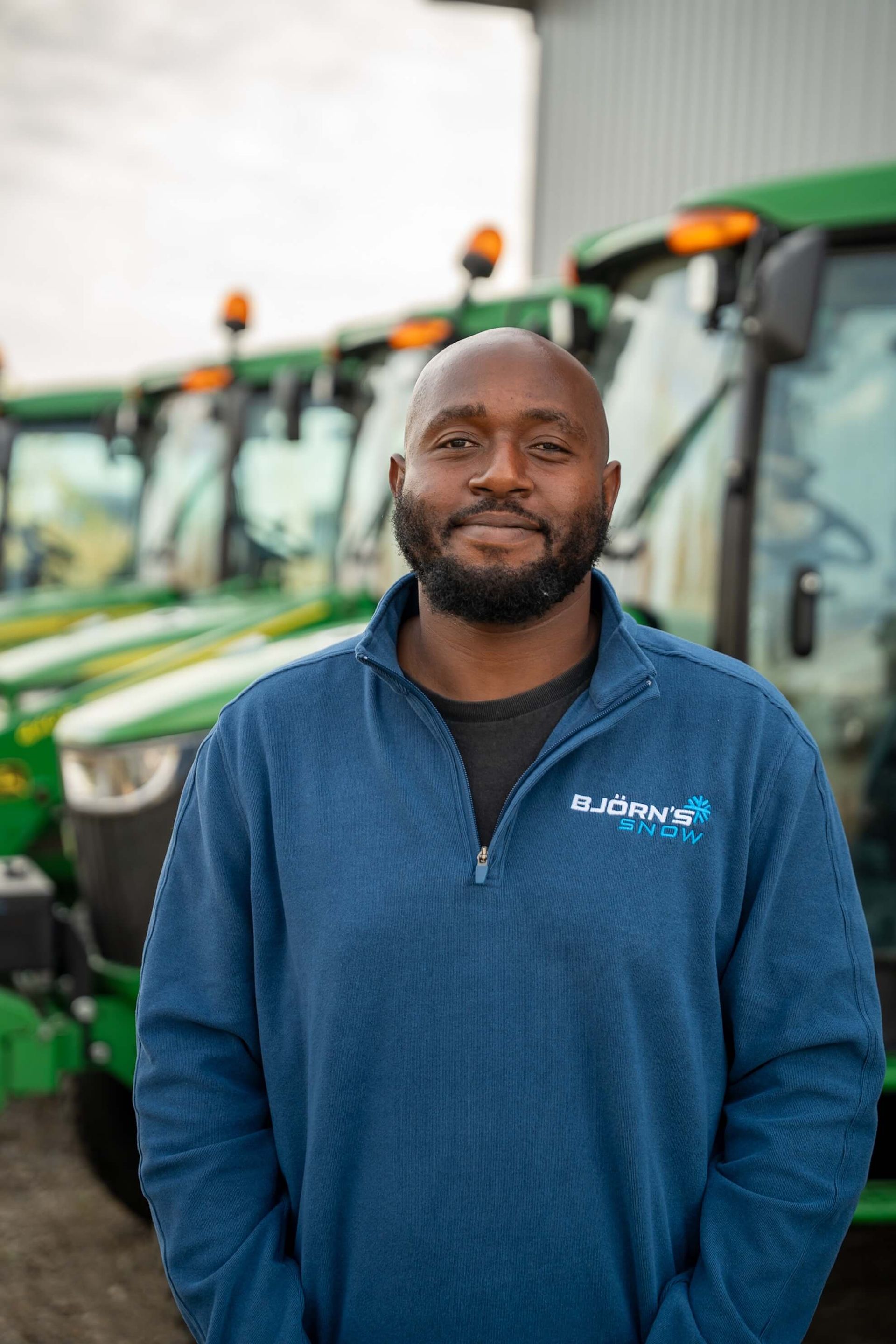 Man in blue sweater smiles in front of green tractors.