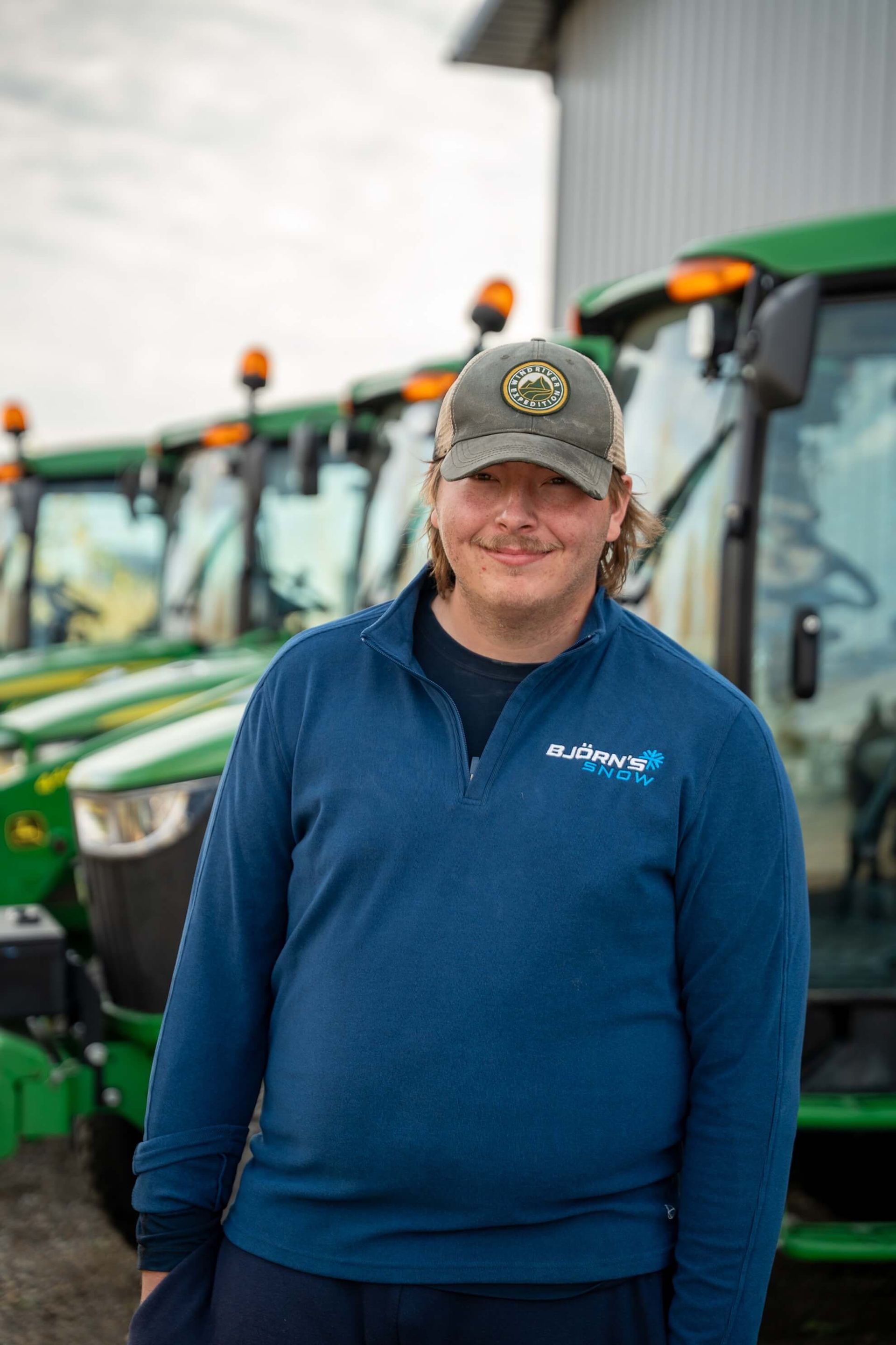 Man in blue shirt and cap stands in front of a row of green tractors.