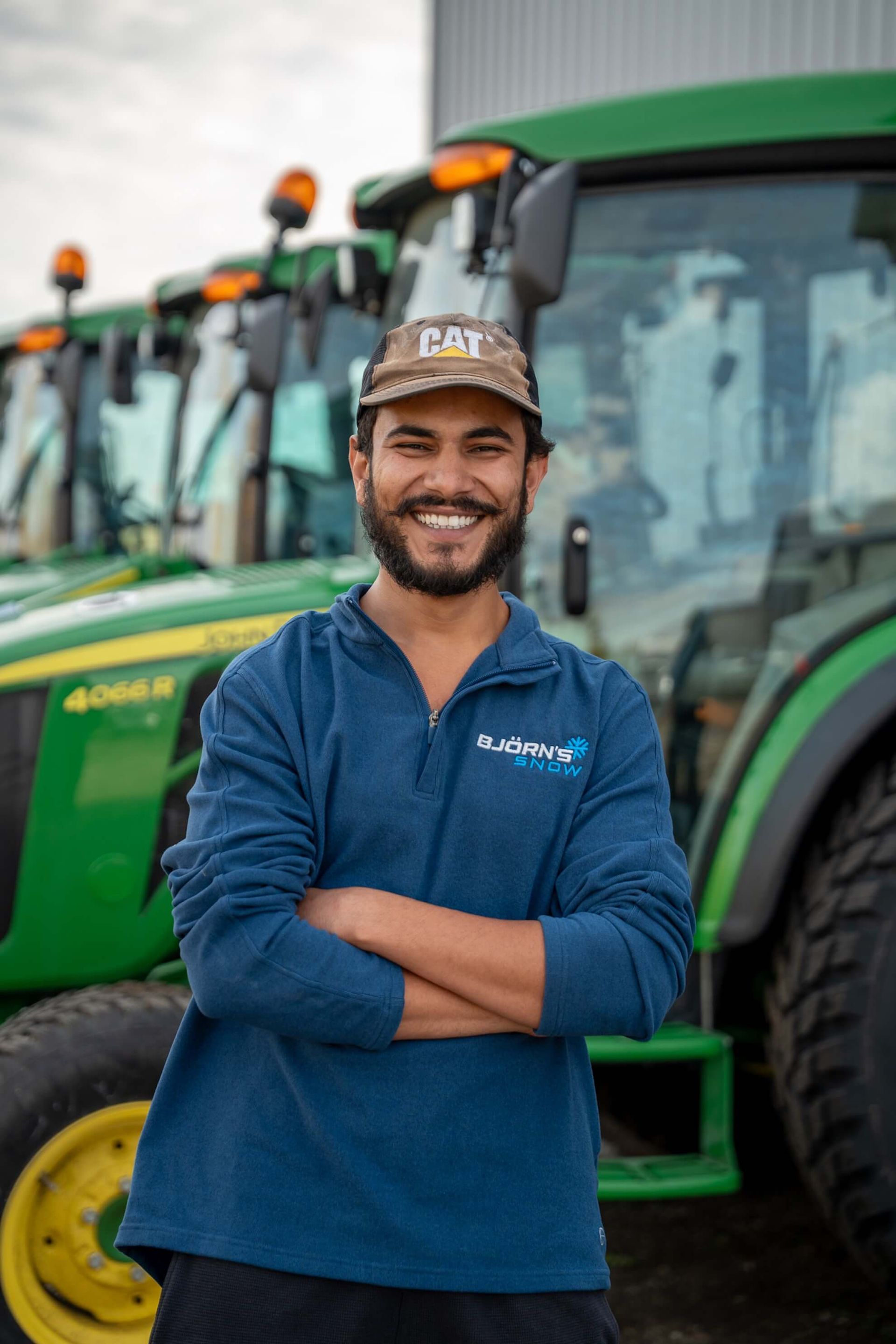 Man smiling, arms crossed in front of green John Deere tractors. He wears a blue shirt and Cat cap.