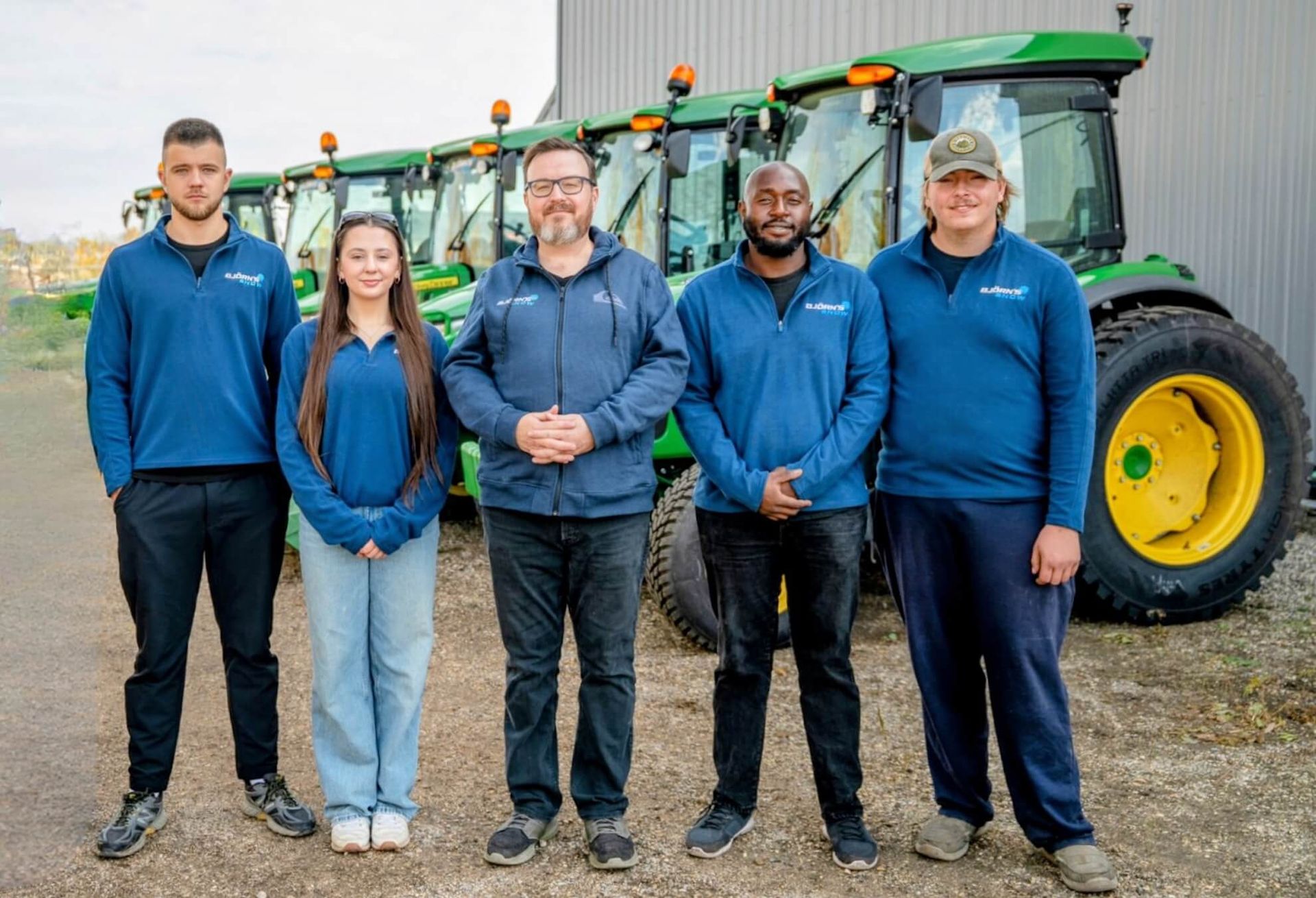 Five people in blue shirts stand in front of green tractors.