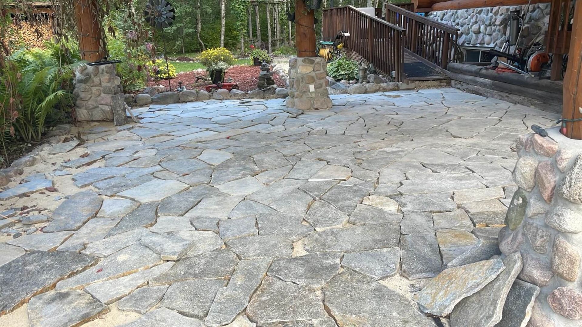 Patio with irregular stone paving, bordered by wooden structures and surrounded by greenery.
