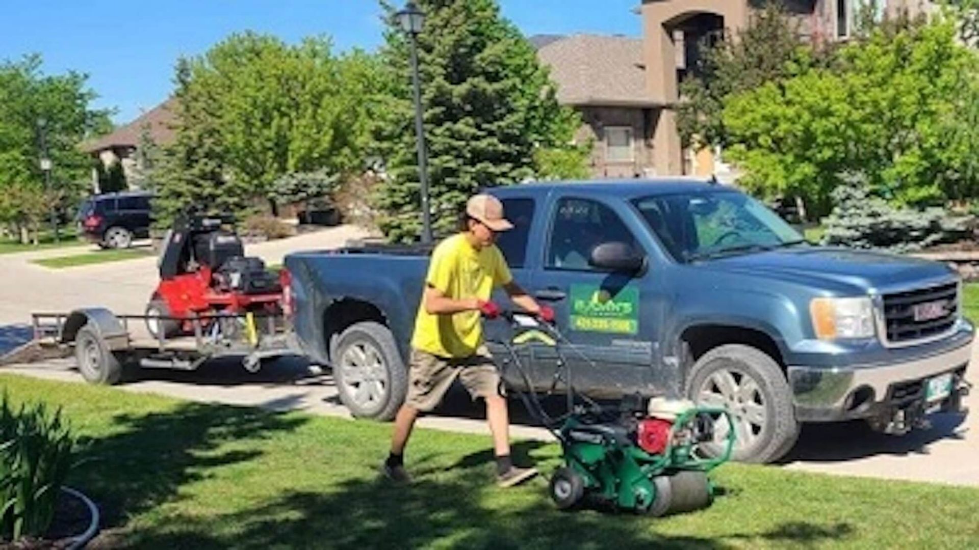 A man mowing a lawn next to a truck with a trailer carrying lawn equipment.