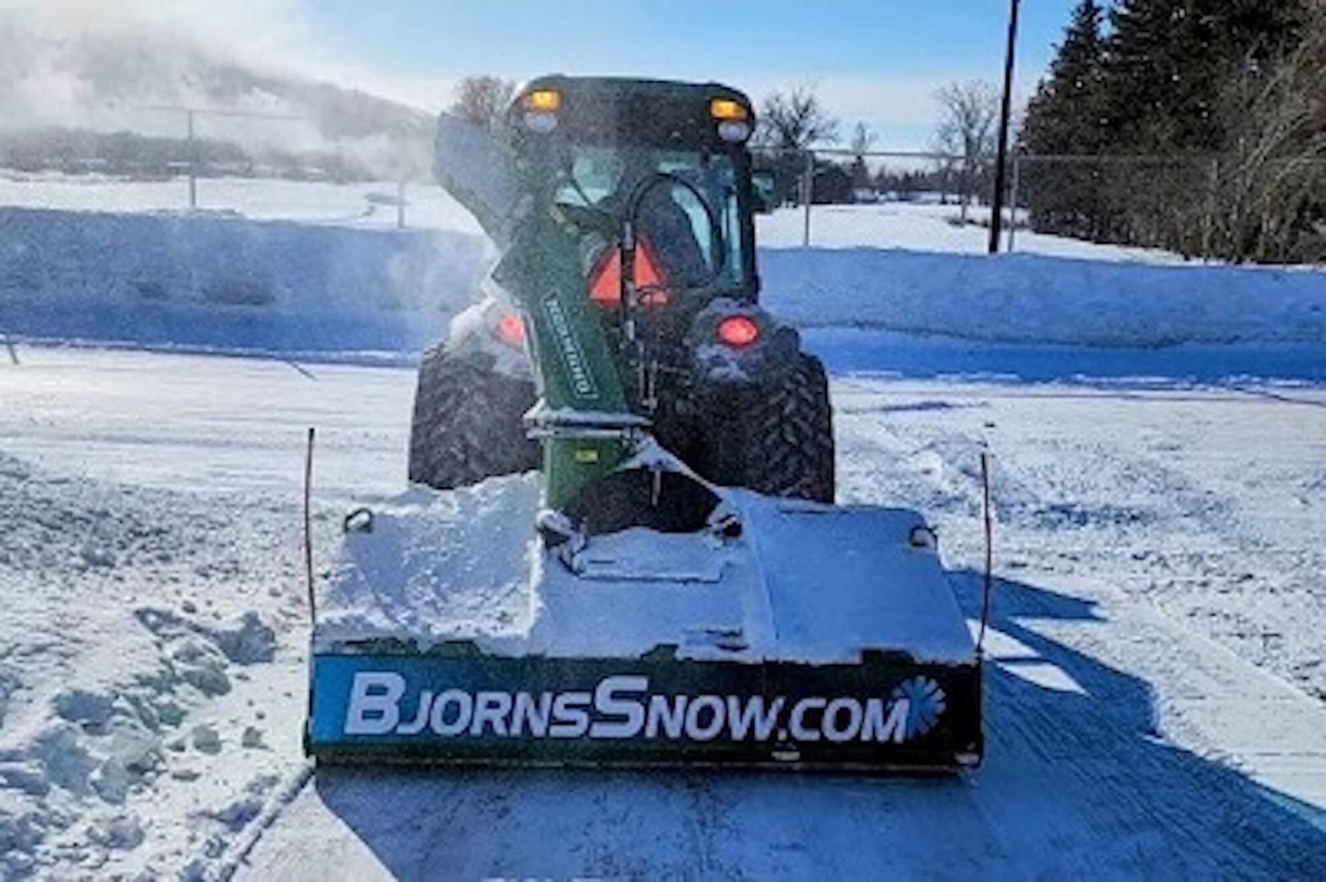 Green tractor with snowblower clearing snow, Bjornessnow.com visible.