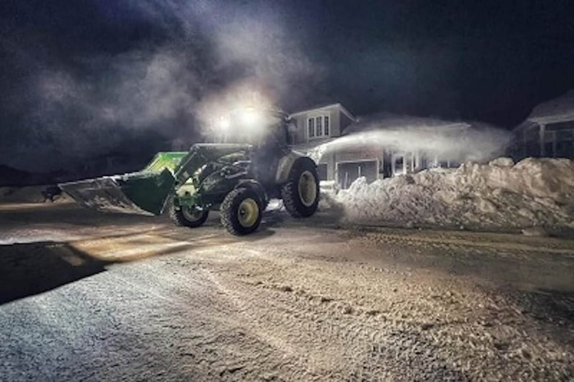 Snow blower clearing a snowy road at night, emitting a cloud of snow; house in the background.