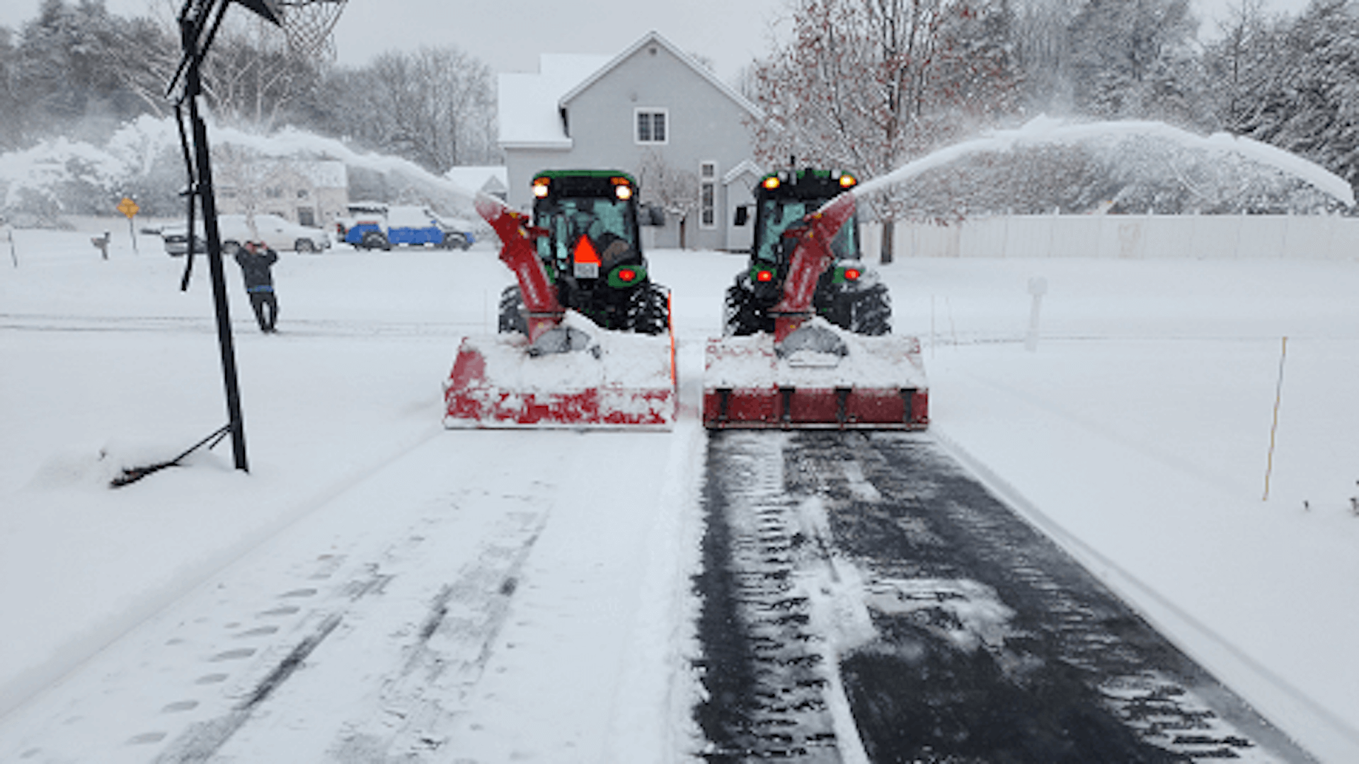 Two tractors with snowblowers clearing a snow-covered road. White snow falling; a house in the background.