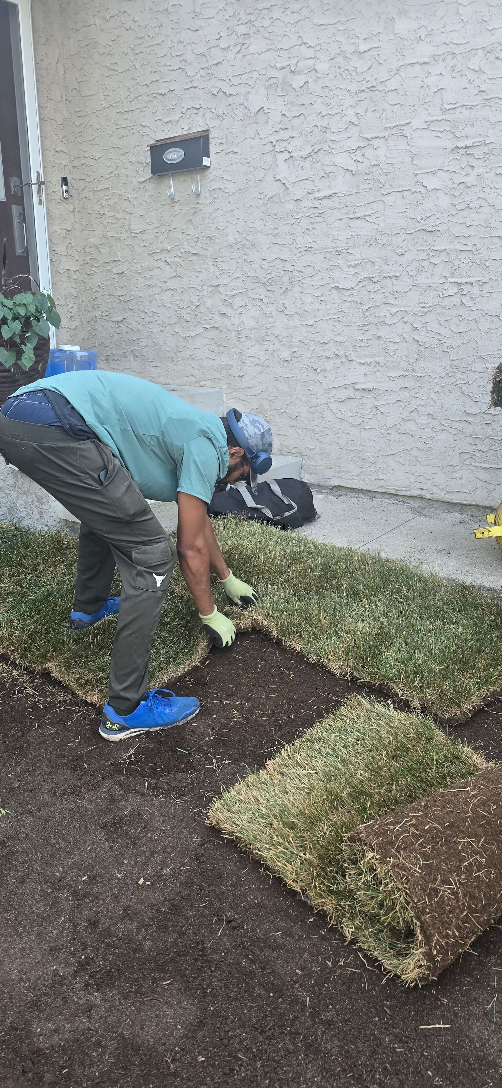 Two people laying sod on the ground. One man wears gloves and a hat. The setting appears outdoors.