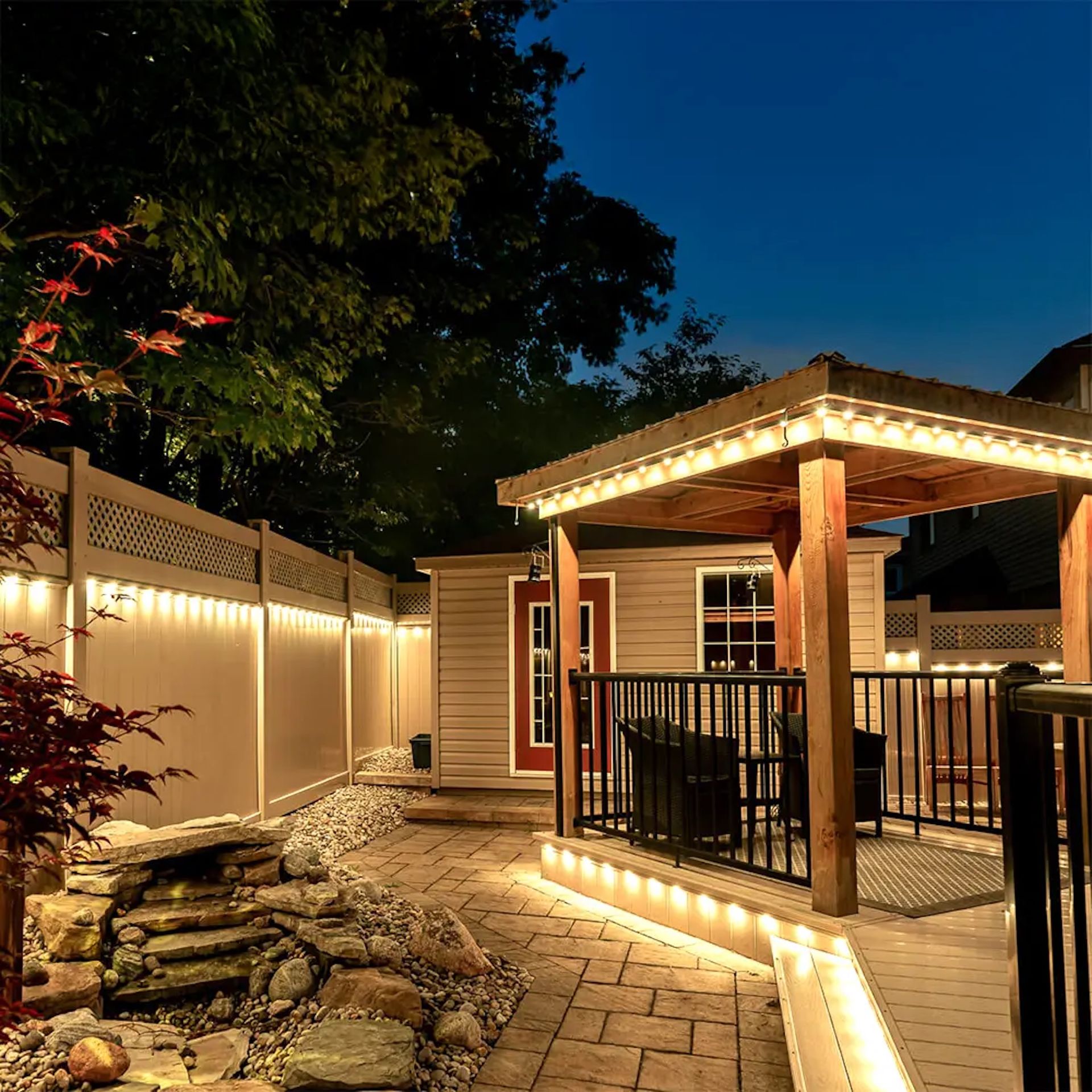 A night view of a landscaped backyard with a wooden pergola, a patio, a stone fountain, and string lights on a fence.