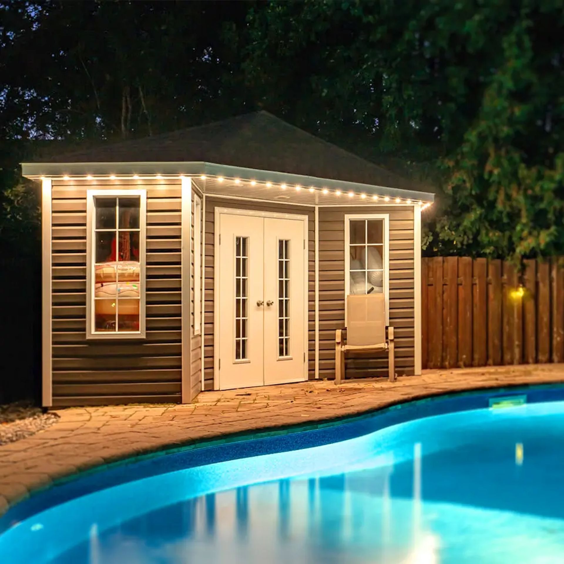 A gray corner shed with white doors and glowing string lights by a lit backyard swimming pool at night.