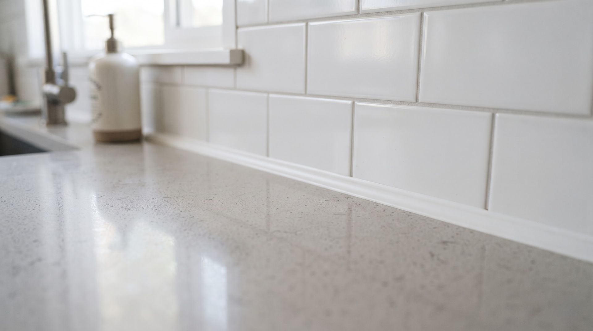 White kitchen countertop beside a glossy white subway tile backsplash and window ledge with soap dispenser.