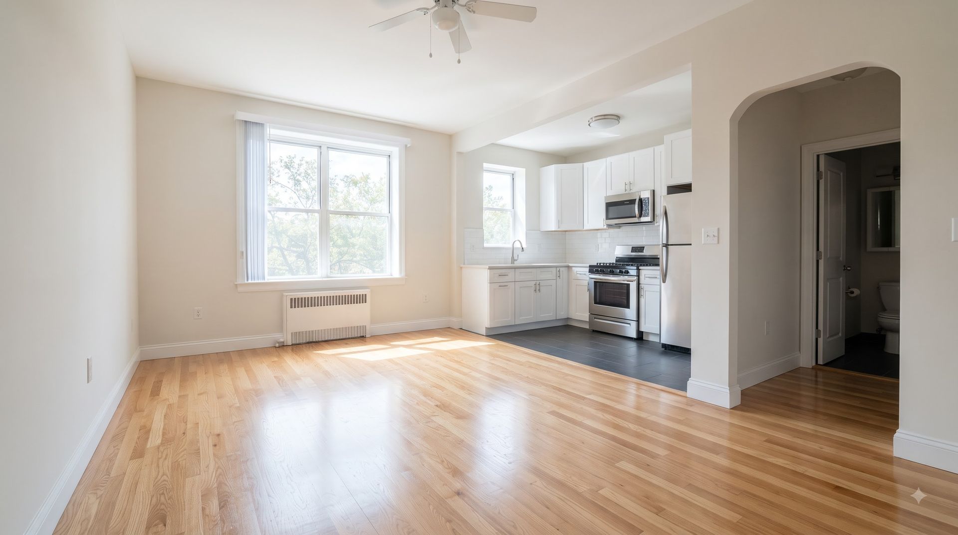 Bright empty living room with hardwood floors, large window, and open kitchen archway