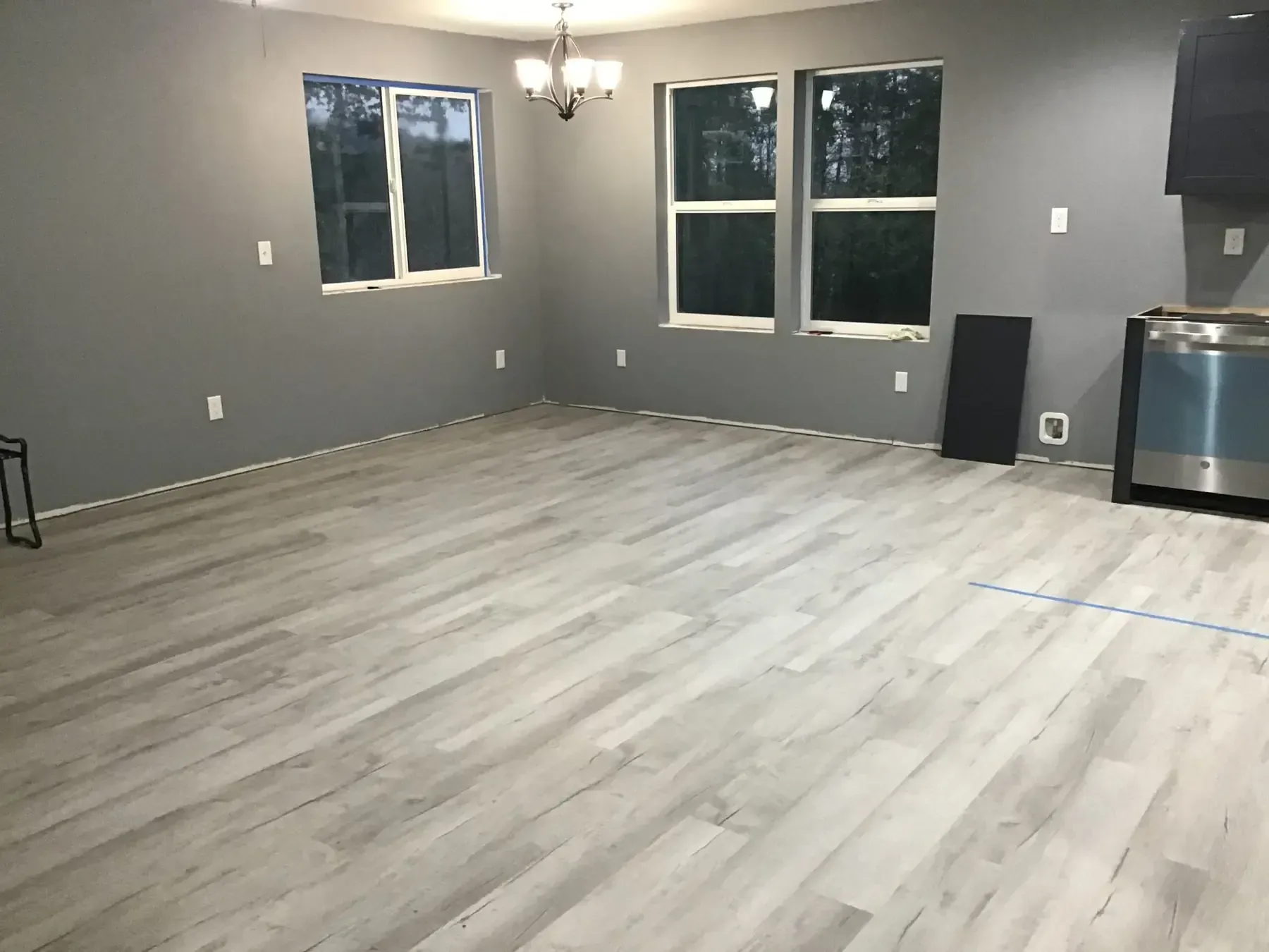 Empty gray room with light wood flooring, windows, chandelier, and a small bar area.