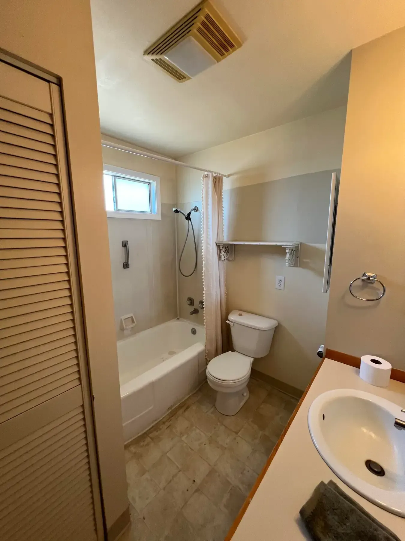 Small bathroom with tub, toilet, sink, and wall shelf; beige tile and louvered closet door.